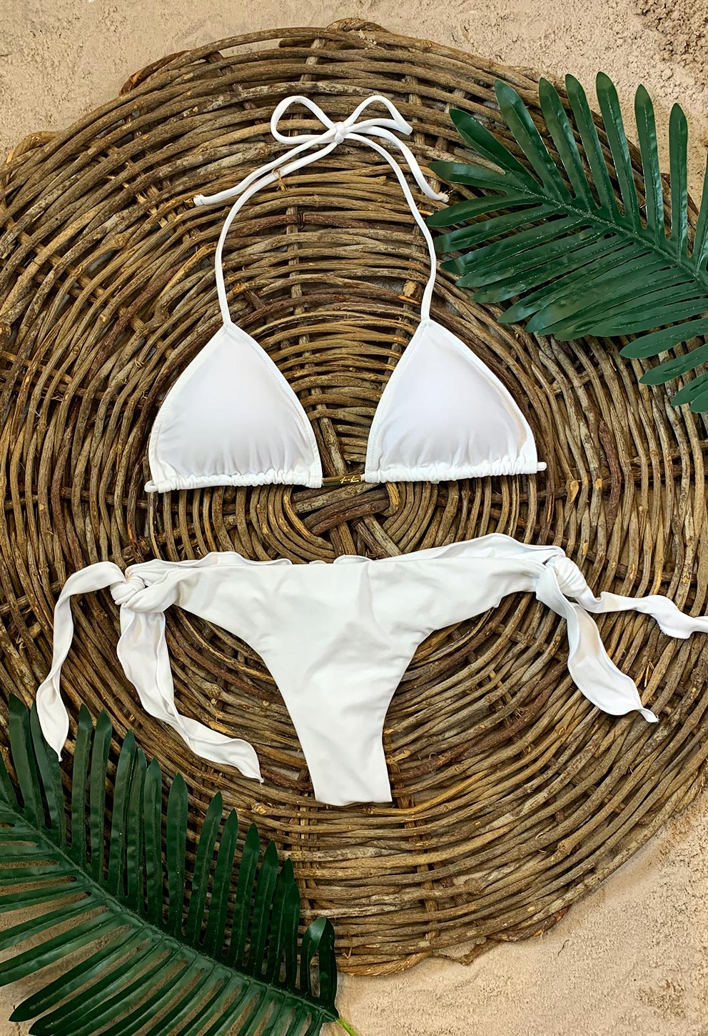 Conjunto de biquíni branco disposto sobre esteira de fibras rústicas e areia. Calcinha de amarração lateral larga com efeito ripple e top cortininha com tiras para amarrar no pescoço; detalhe drapeado
