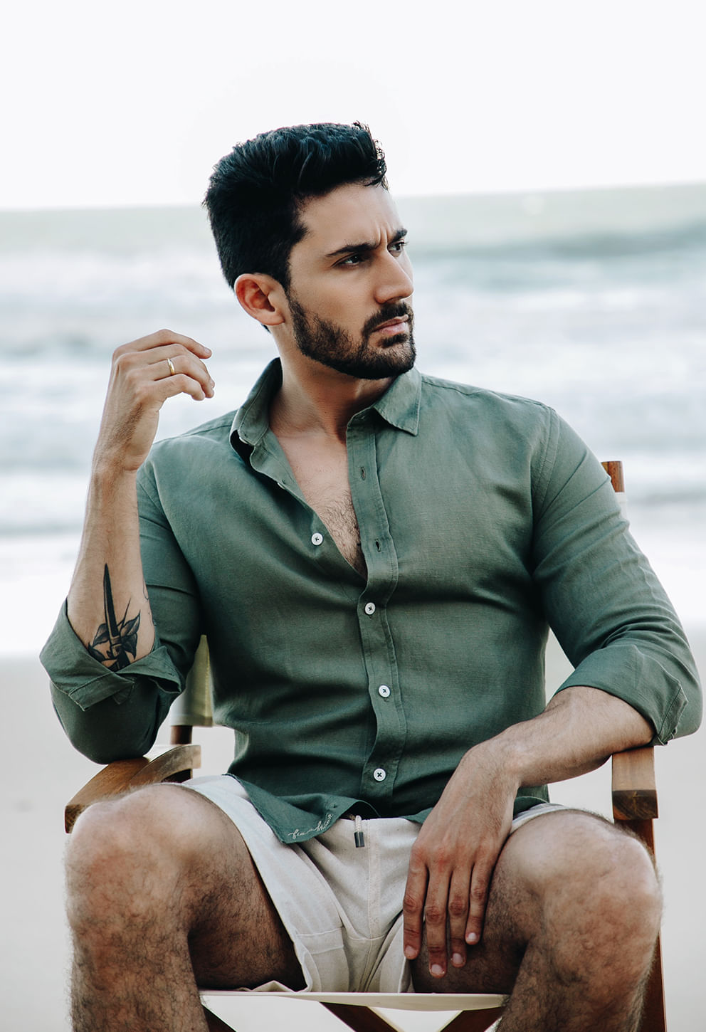 Homem sentado na praia vestindo camisa manga longa verde militar, tecido com aparência de linho e barra com assinatura bordada