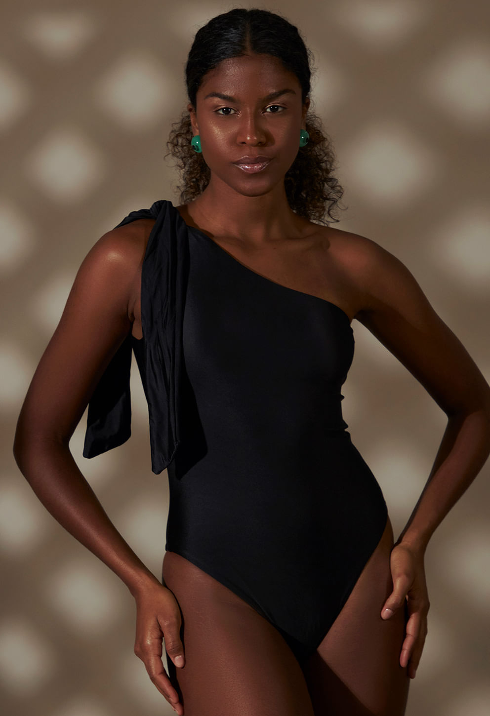 Mulher morena com cabelo cacheado preso, usando body preto de um ombro só com nó largo no ombro esquerdo, posicionado de frente com leve inclinação para o lado. Body liso acetinado, corte alto na per