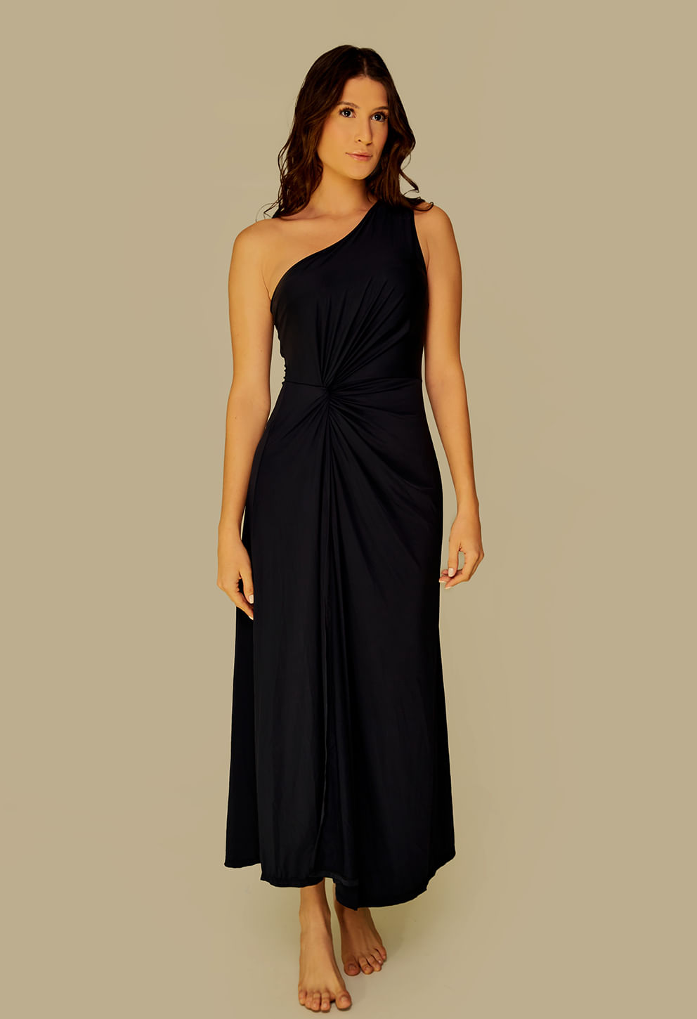 Vestido longo preto com ombro único, drapeado na cintura e fenda lateral, modelo em pé com fundo bege.