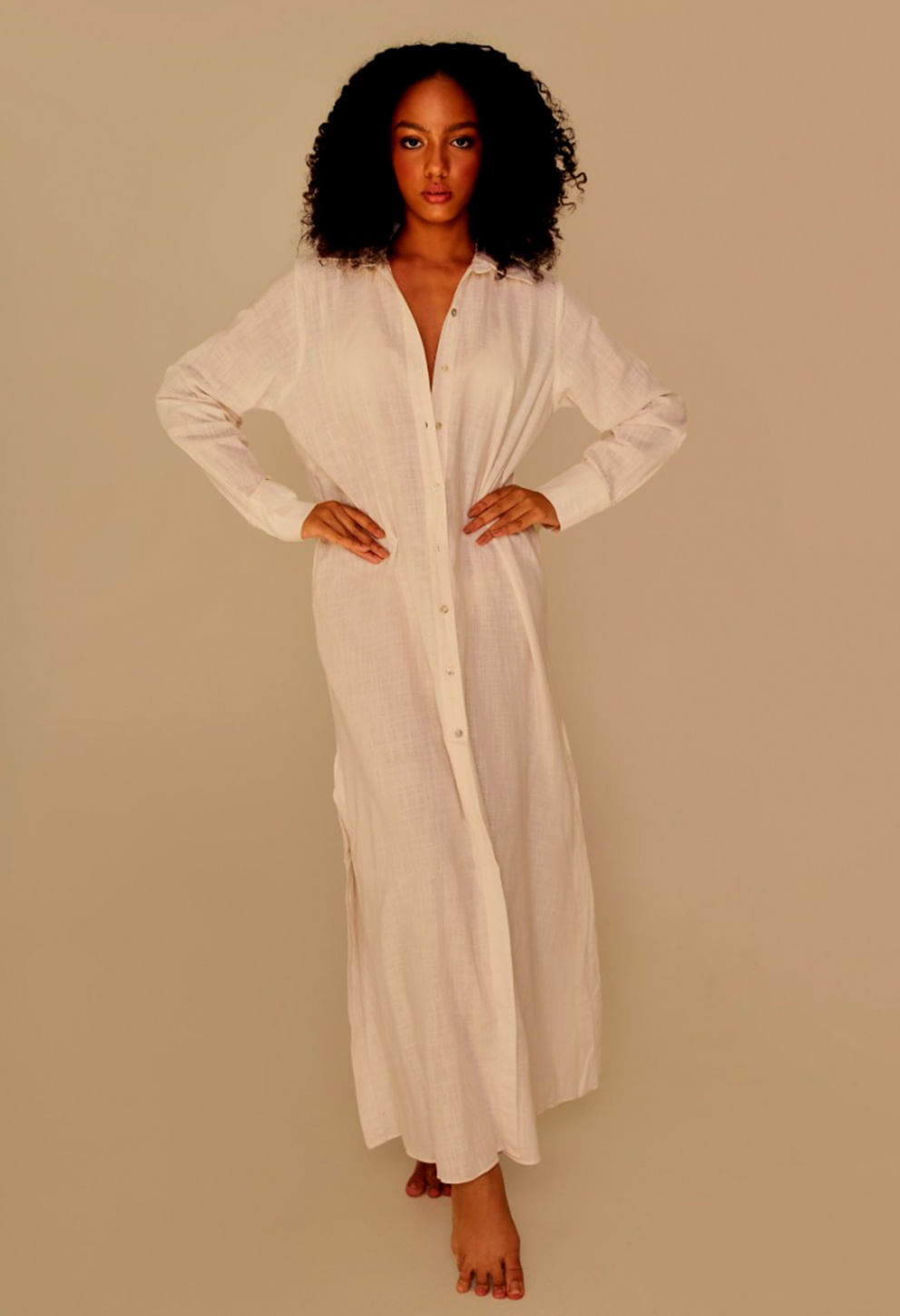 Mulher morena de cabelo cacheado usando chemise longa de cambraia off white, com botões frontais e mangas longas, em fundo bege