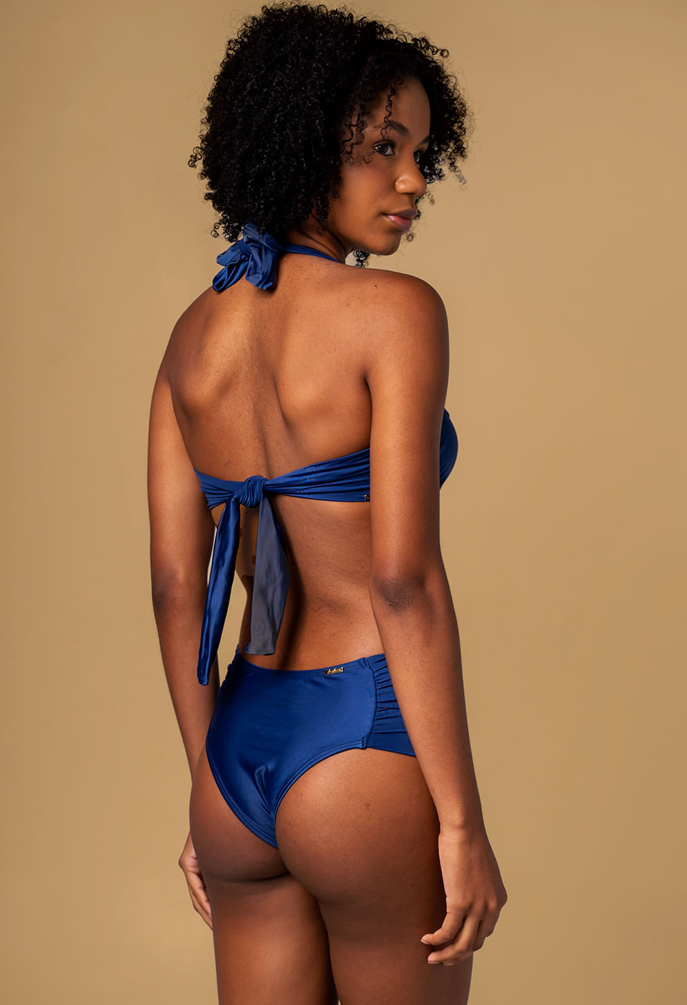 Modelo feminina mostrando amarração do top em laço no meio das costas, biquíni azul marinho acetinado