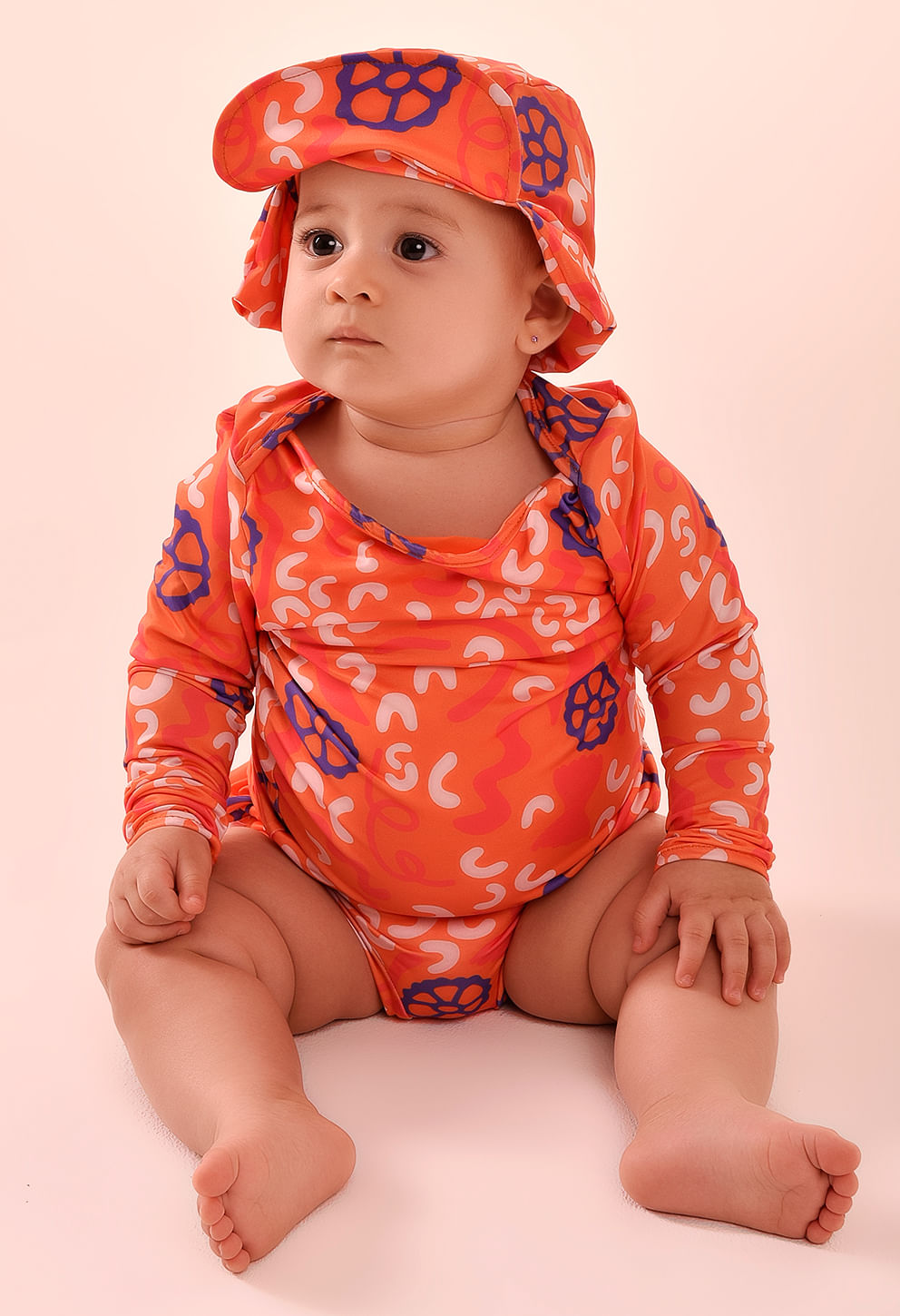 Bebê sentado vestindo conjunto laranja com estampa e chapéu combinando; destaque na calcinha baby com babado duplo traseiro.