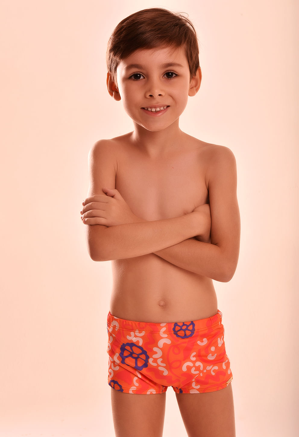 Imagem frontal de um menino sorridente usando uma sunga short infantil com estampa vibrante predominantemente em laranja, com detalhes em azul e branco.