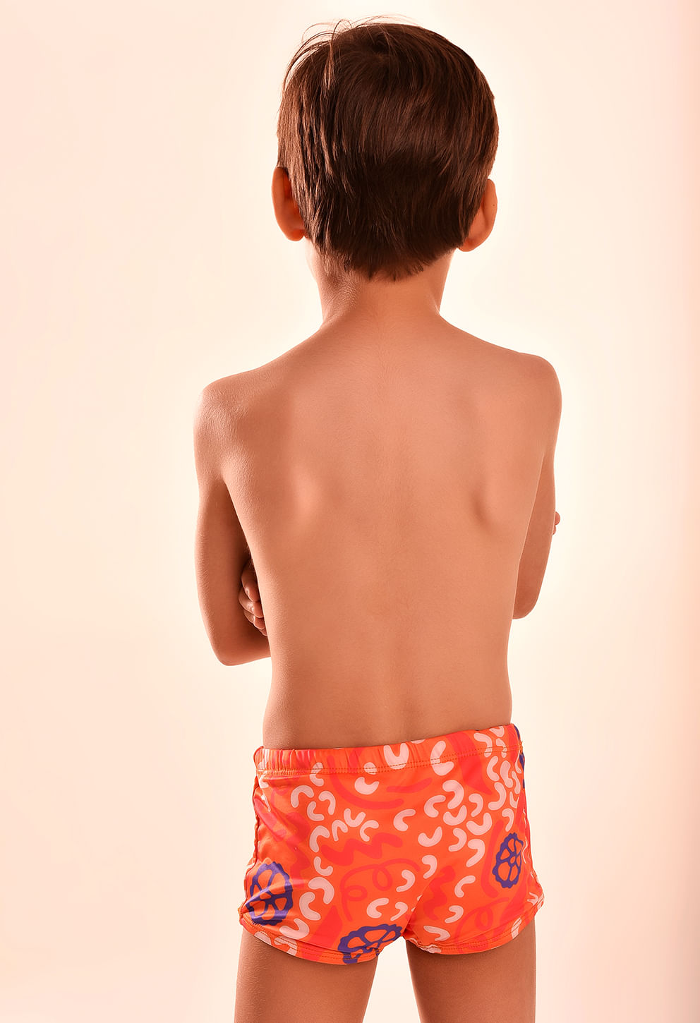Imagem traseira de um menino de costas usando uma sunga short infantil estampada em laranja vibrante com detalhes brancos e azuis.