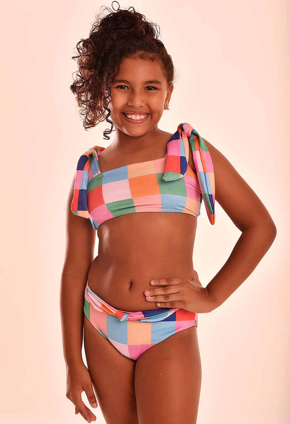 Menina modelo infantil usando biquíni tipo top e calcinha com estampa xadrez colorida (quadrados em rosa, laranja, azul, verde e salmão). Top reto com alças largas que formam laços nos ombros e amarra