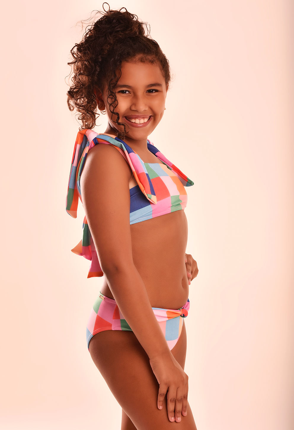 Perfil direito da menina modelo infantil usando o biquíni xadrez multicolor com alça larga e laço nas costas, mostrando conforto e estilo.