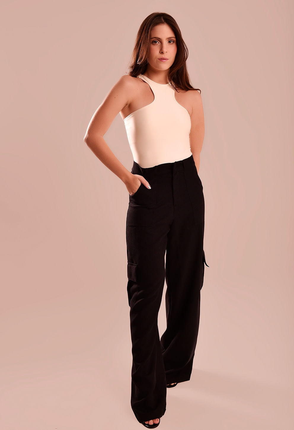 Calça pantalona preta frontal com bolsos cargo e regata creme, modelo com mão no bolso, fundo bege rosado.