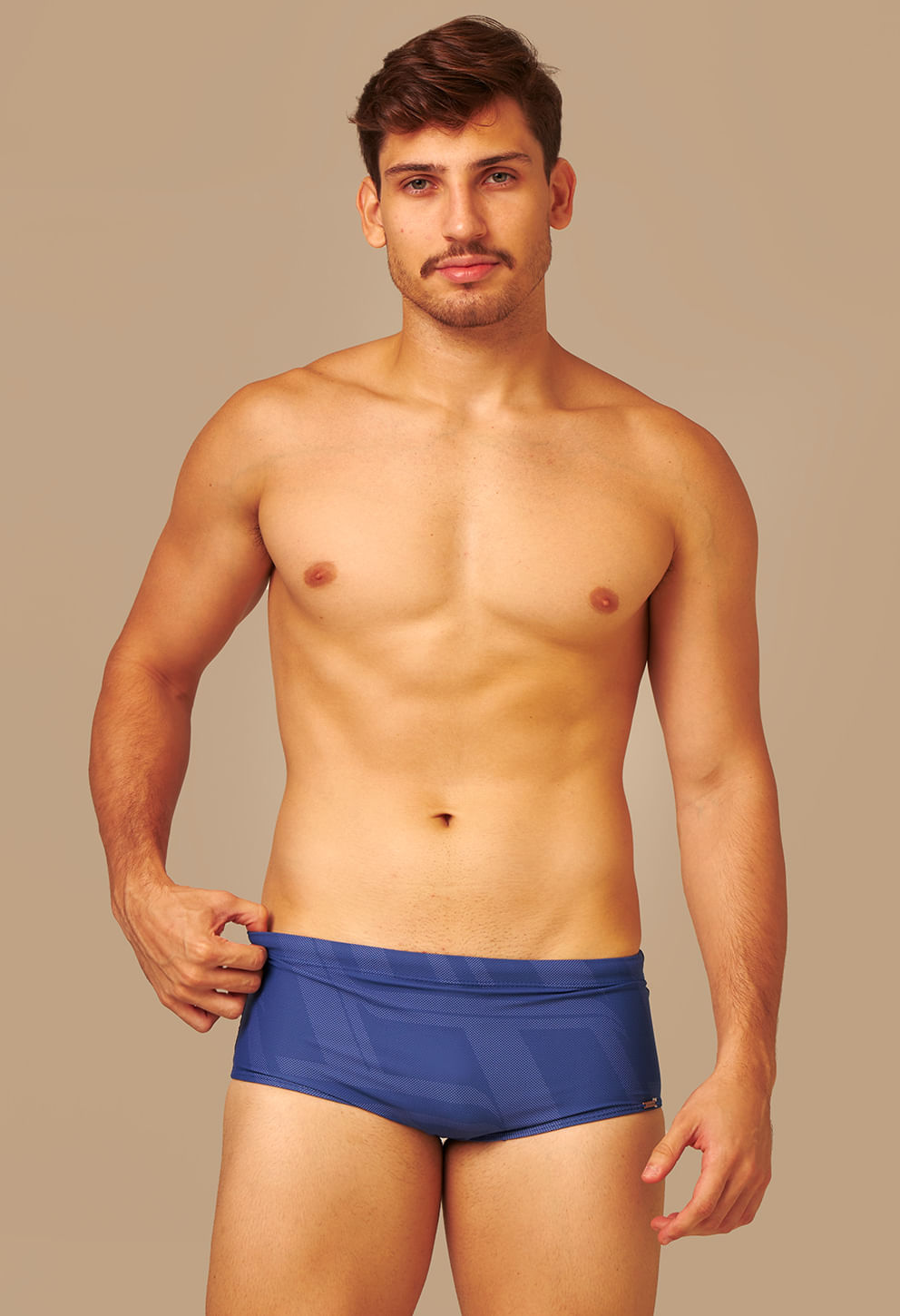 Homem com corpo atlético usa sunga short adulto azul com estampa geométrica em tons de azul, posicionado de frente contra um fundo bege neutro, segurando a cintura da sunga com a mão esquerda, a sunga