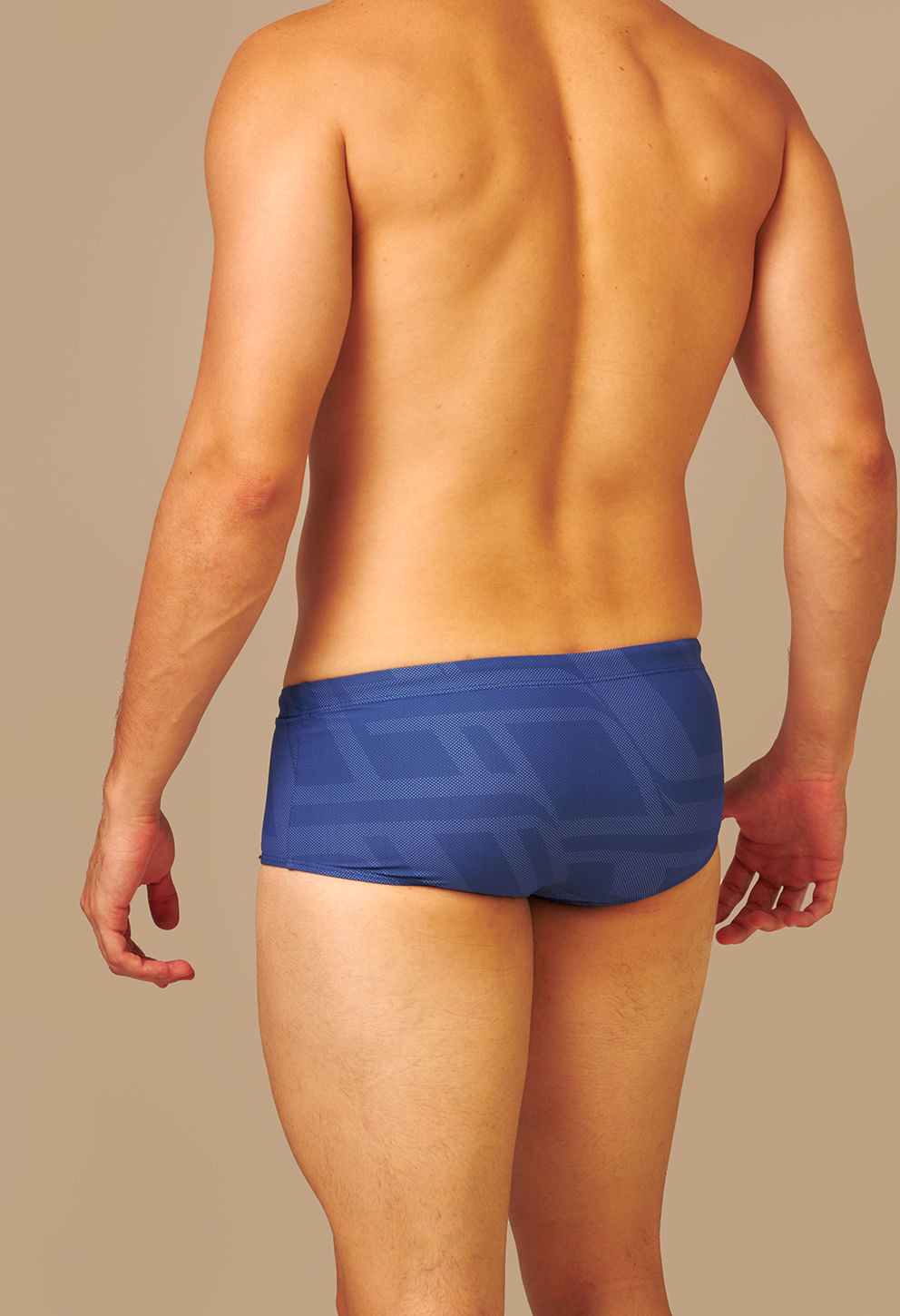 Parte traseira de homem atlético vestindo sunga short adulto azul com estampa geométrica em tons de azul, destacando a modelagem reta e cobertura média, contra fundo bege neutro