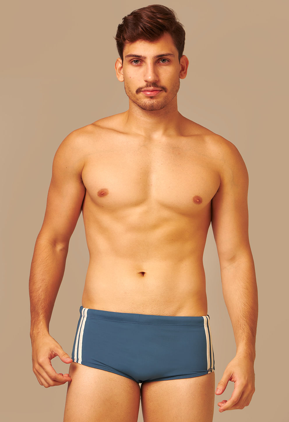 Homem adulto usando sunga short na cor petróleo, com duas listras verticais brancas nas laterais. A imagem mostra o modelo de frente, com o rosto e torso visíveis, em fundo bege neutro. A sunga tem c