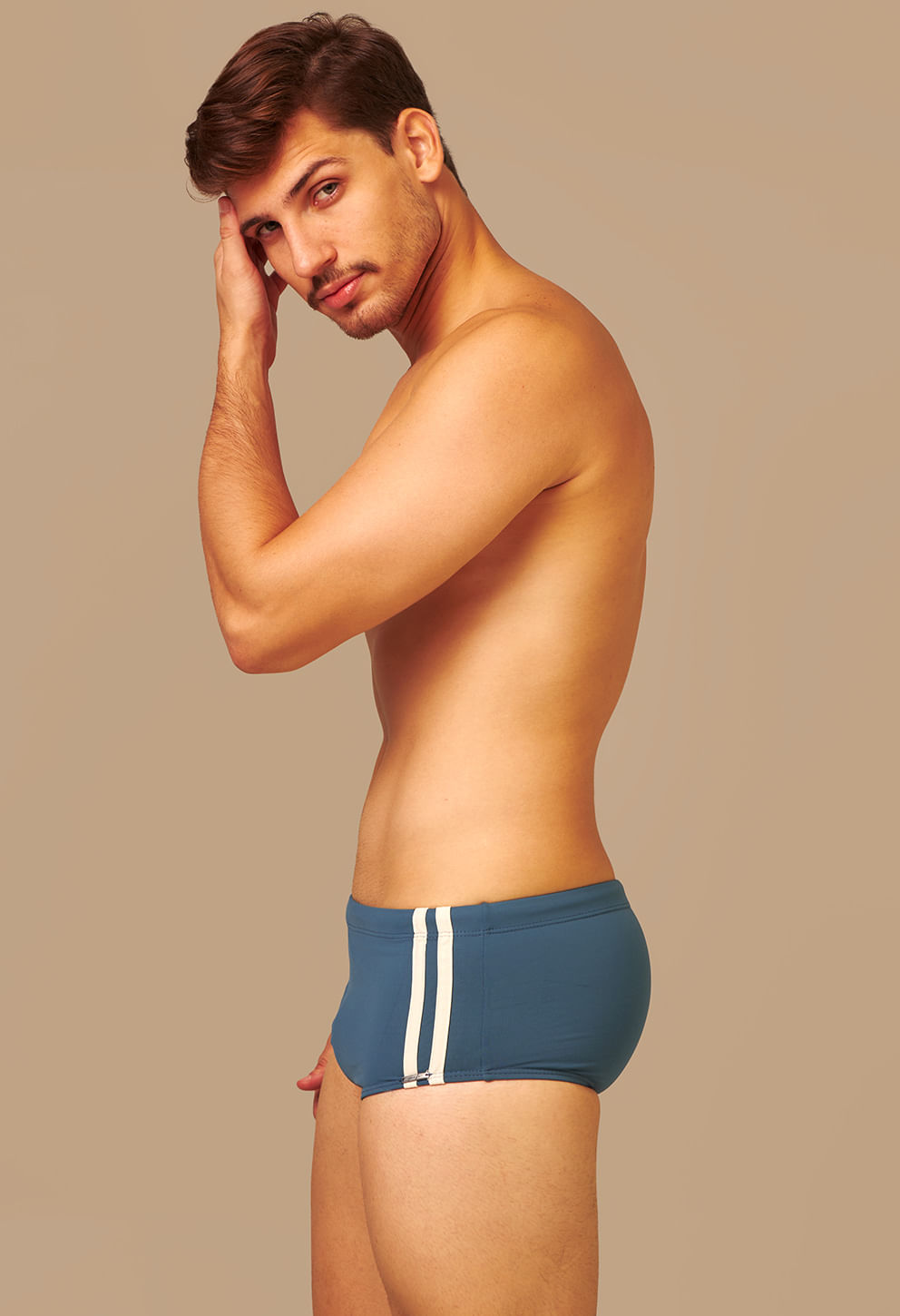 Modelo masculino exibindo sunga short petróleo com duas listras brancas verticais na lateral direita. Foto capturada de perfil, focando no ajuste e caimento da peça, com fundo bege liso que destaca o