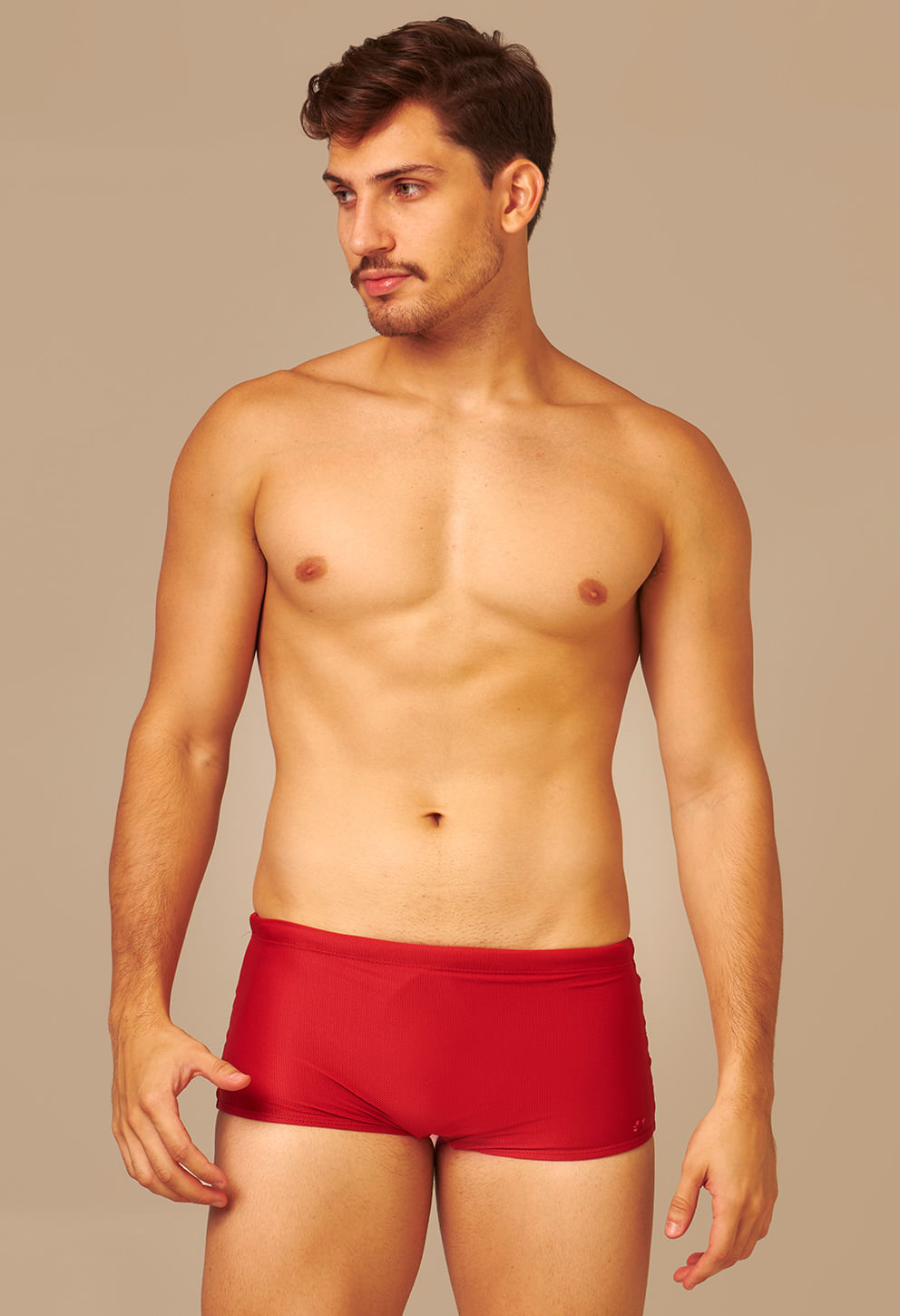 Homem modelo usando sunga short adulto vermelha vibrante com corte justo e tecido texturizado em fundo bege neutro.