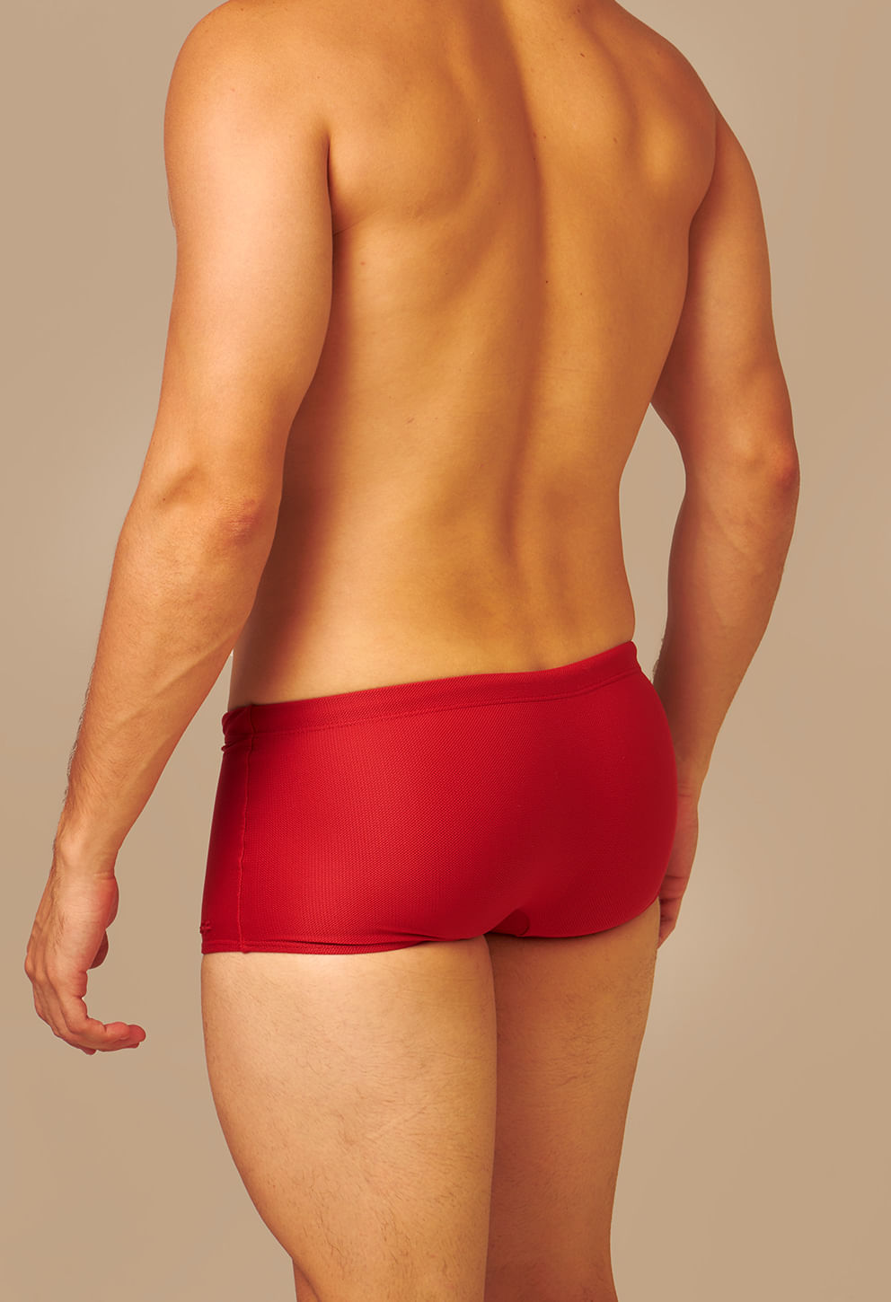 Modelo de costas usando sunga short vermelho vibrante com corte justo e tecido texturizado em fundo neutro.