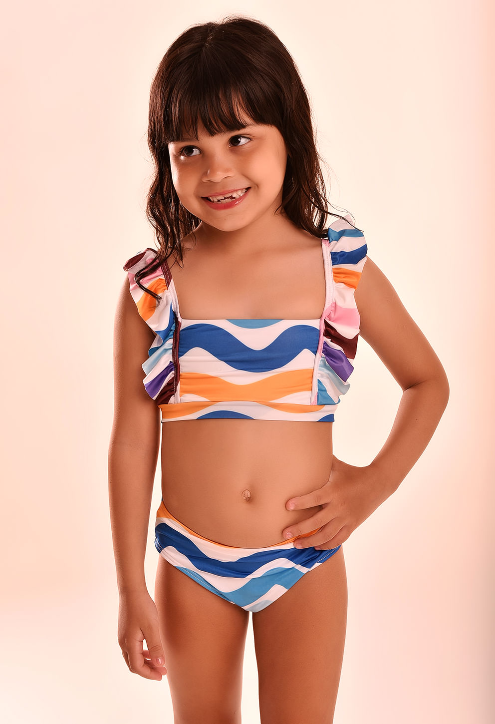 Menina sorridente usando biquíni infantil Ariel Chuá com estampa em ondas coloridas predominando azul, laranja, rosa, roxo, branco e marrom.