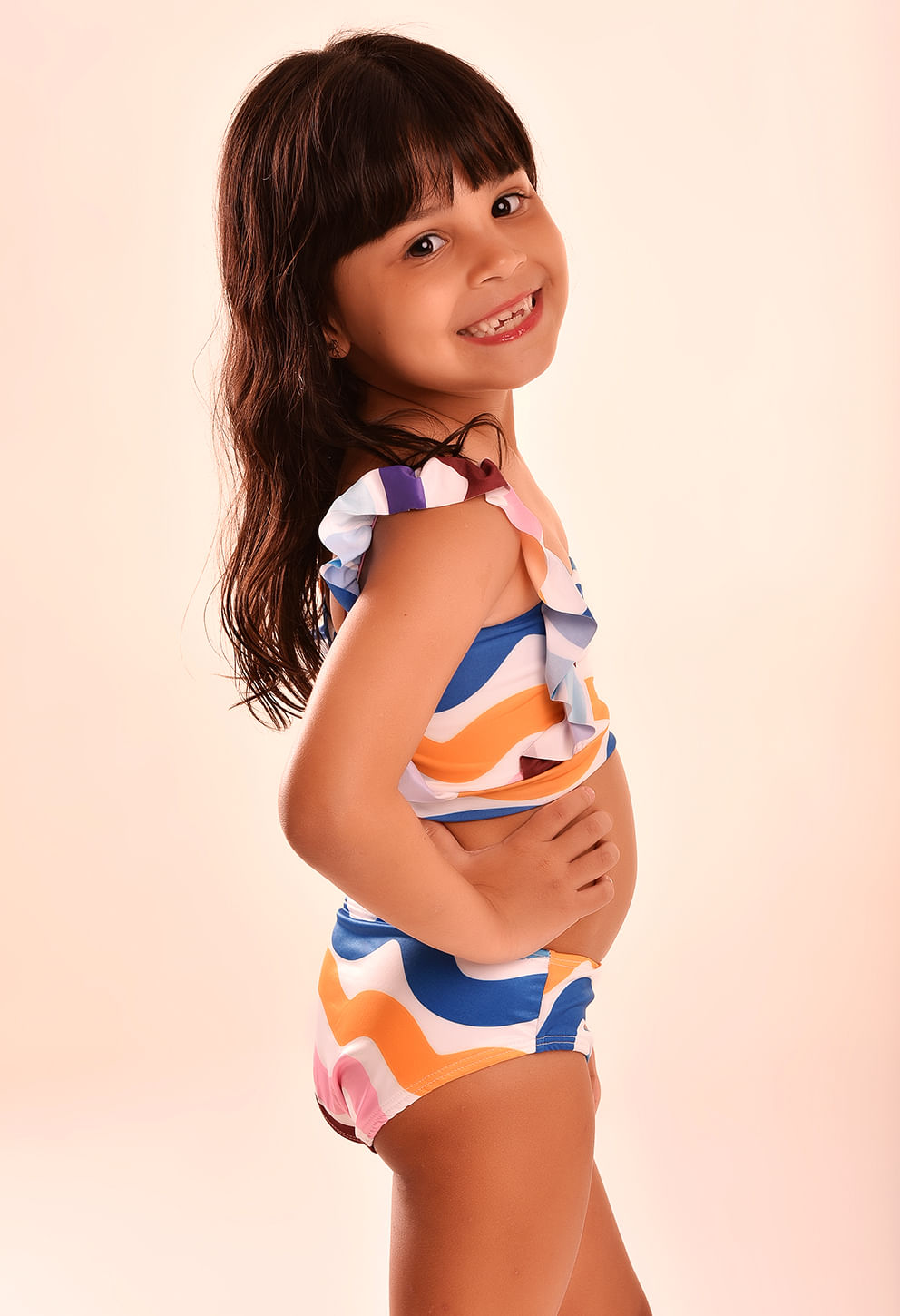 Menina sorrindo de lado vestindo biquíni Ariel Chuá infantil com estampa ondulada colorida em azul, laranja, rosa, roxo, branco e marrom.