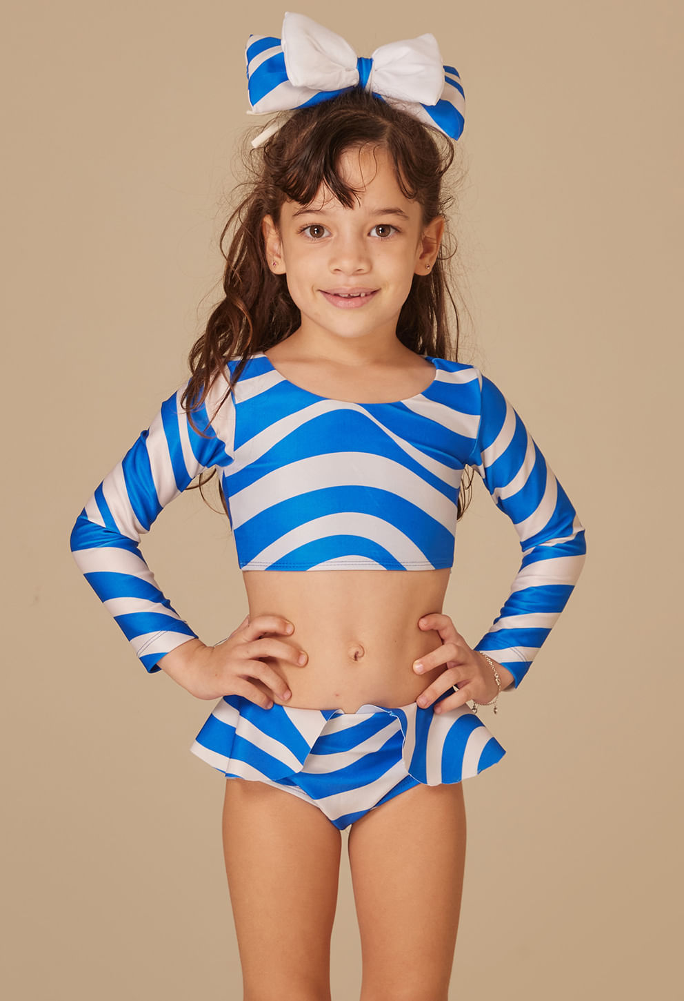 Menina vestindo biquíni infantil Dora Maré azul e branco, manga longa e babado na cintura, laço no cabelo, vista frontal.