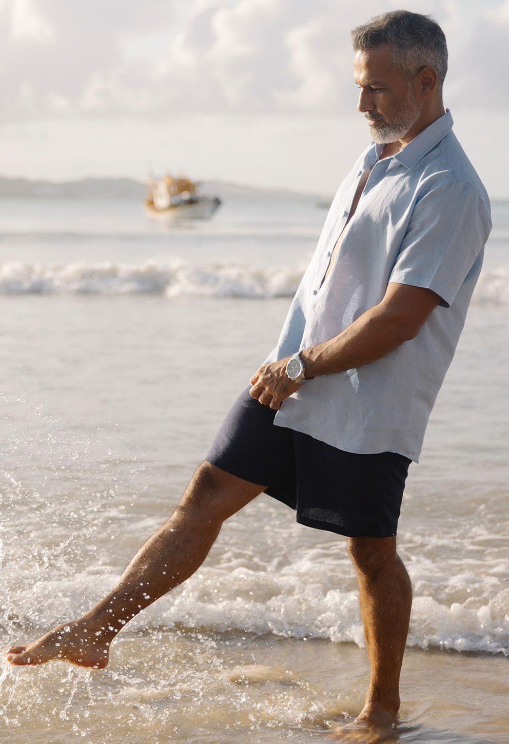 Modelo na praia usando camisa azul claro de linho slim manga curta, combinada com bermuda preta