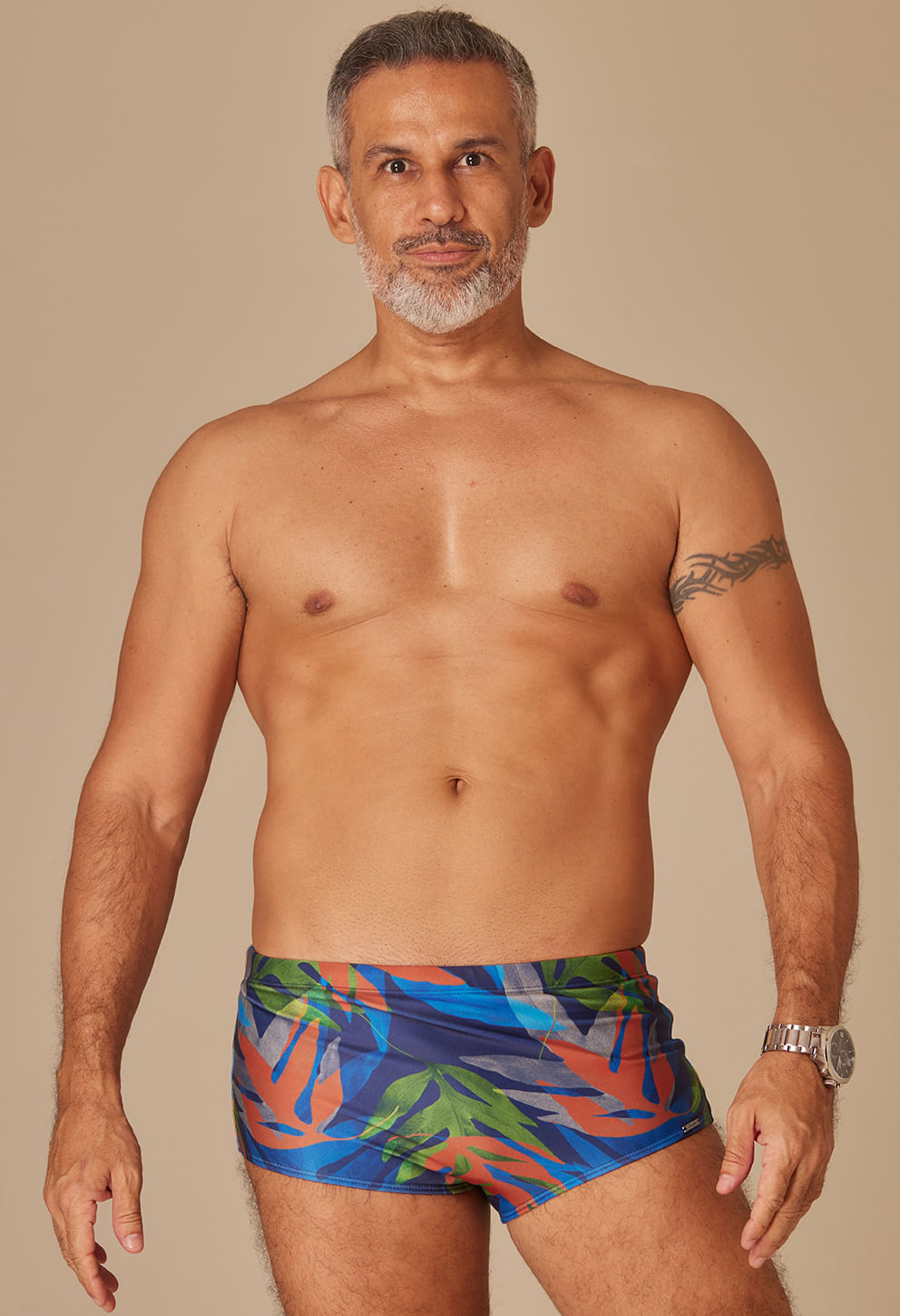 Homem adulto com cabelo grisalho e barba branca, vestindo sunga sungão com estampa de folhas em tons de azul, verde, laranja e cinza.