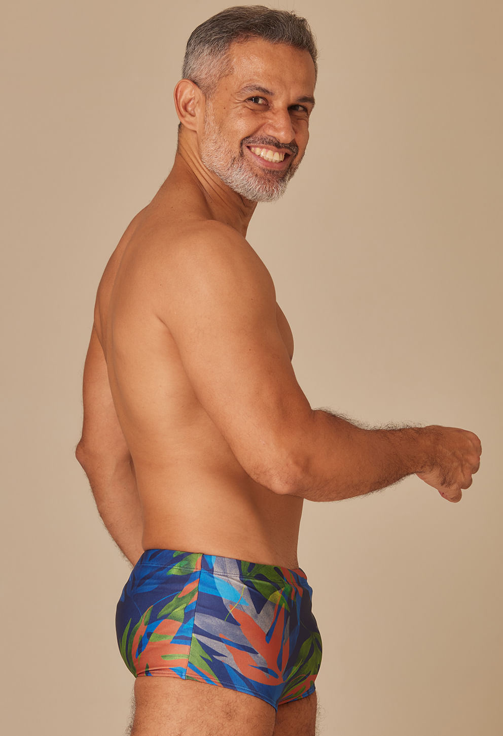 Homem adulto sorridente de perfil usando sunga sungão estampada com folhas em azul, verde, laranja e cinza.