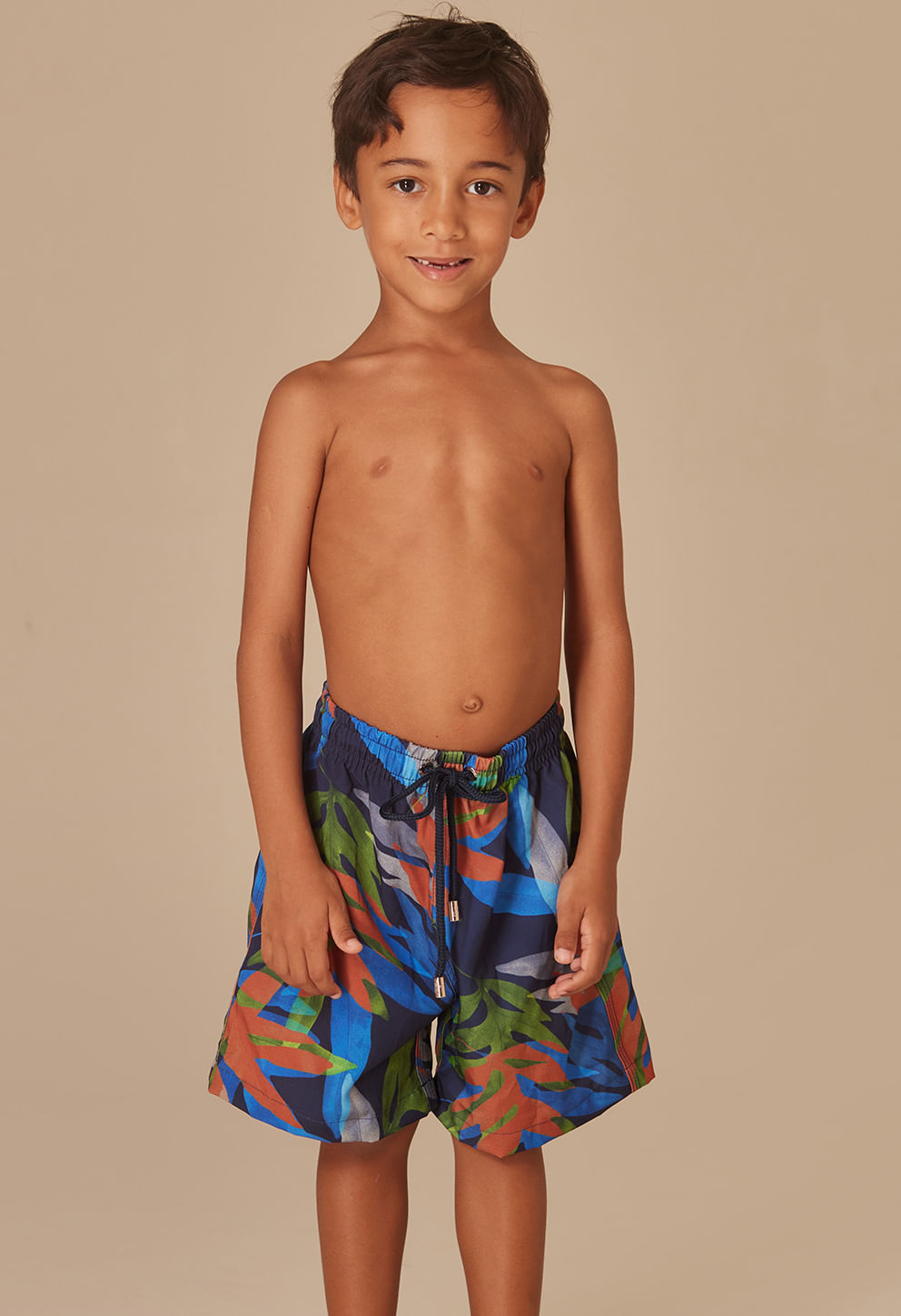 Imagem frontal de um menino criança vestindo uma bermuda infantil com estampa colorida de folhas em tons predominantes de azul, verde, laranja e cinza. A bermuda tem cós elástico com cordão preto para