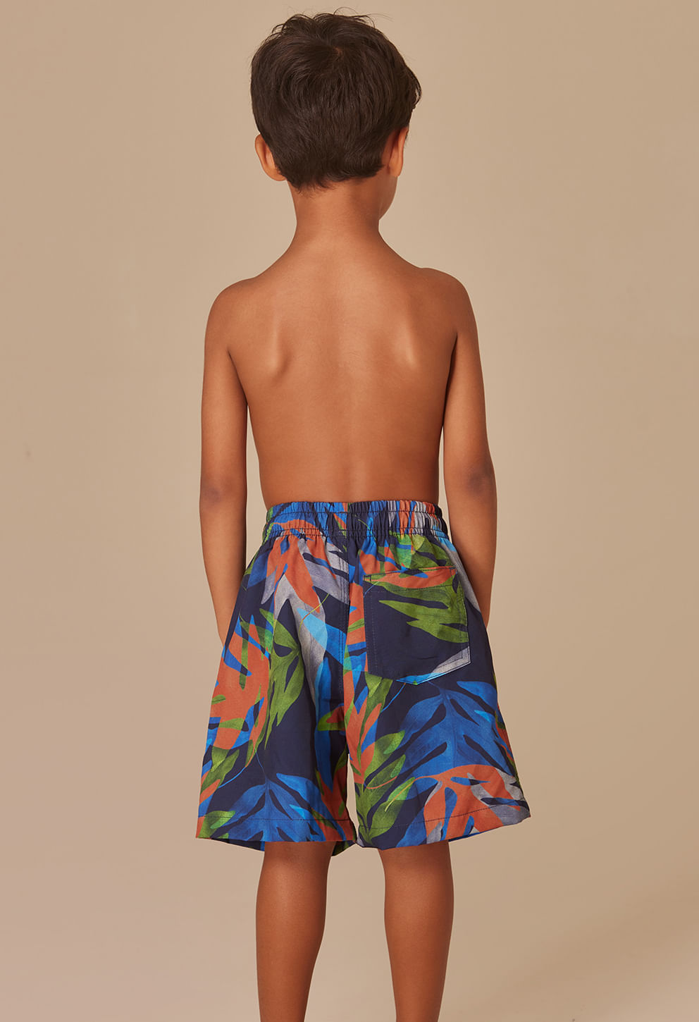Visão traseira de menino vestindo bermuda infantil com estampa colorida de folhas nas cores azul, verde, laranja e cinza, com bolso traseiro direito e cós elástico com cordão para ajuste.