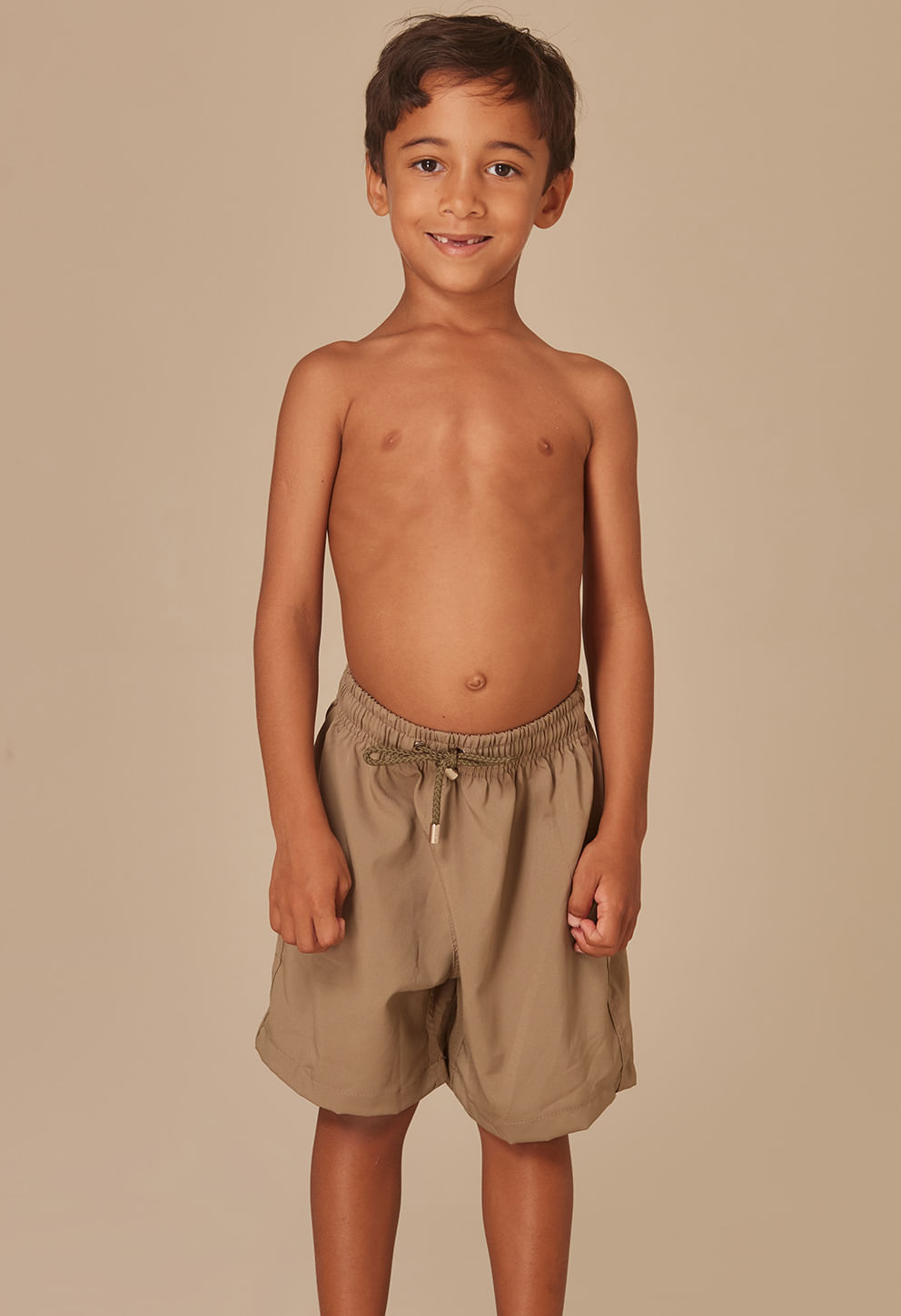 Menino sorridente usando bermuda bege com cós elástico e cordão verde para ajuste, tecido leve e bolsos laterais.