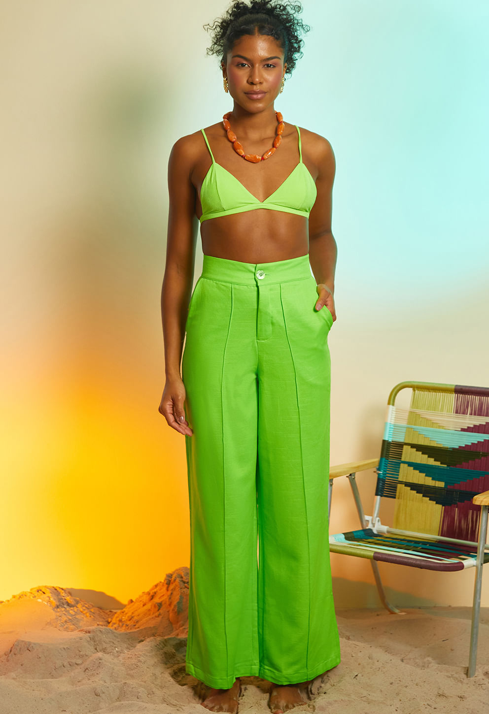 Modelo em pé sobre areia usando calça pantalona verde claro de cintura alta com pregas e bolsos, combinada com top verde e acessórios.