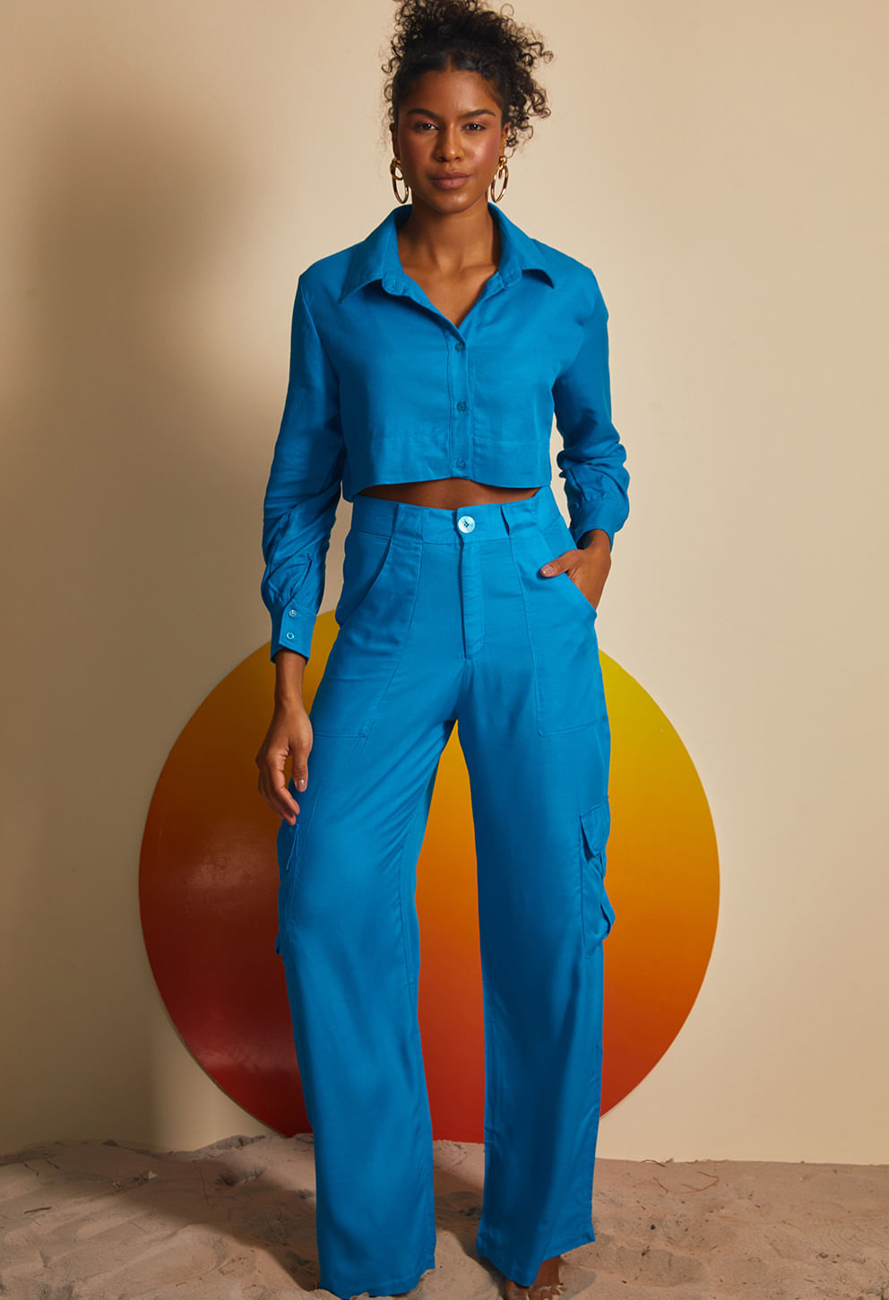 Mulher vestindo camisa cropped azul vibrante de mangas longas com gola e botões frontais, combinada com calça cargo azul