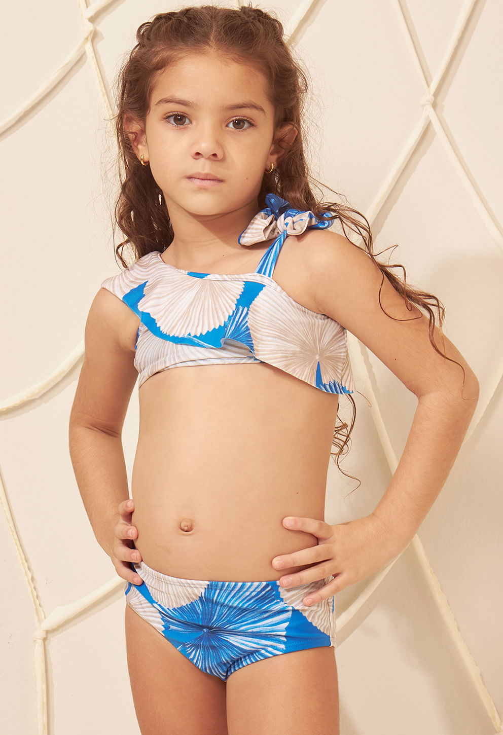 Menina posando de frente com biquíni ombro só infantil estampado em azul, bege e branco, com babado assimétrico e laço no ombro.