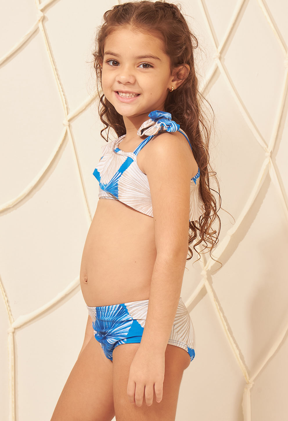 Menina sorrindo de lado usando biquíni ombro só infantil estampado em azul, bege e branco, com babado e laço no ombro.