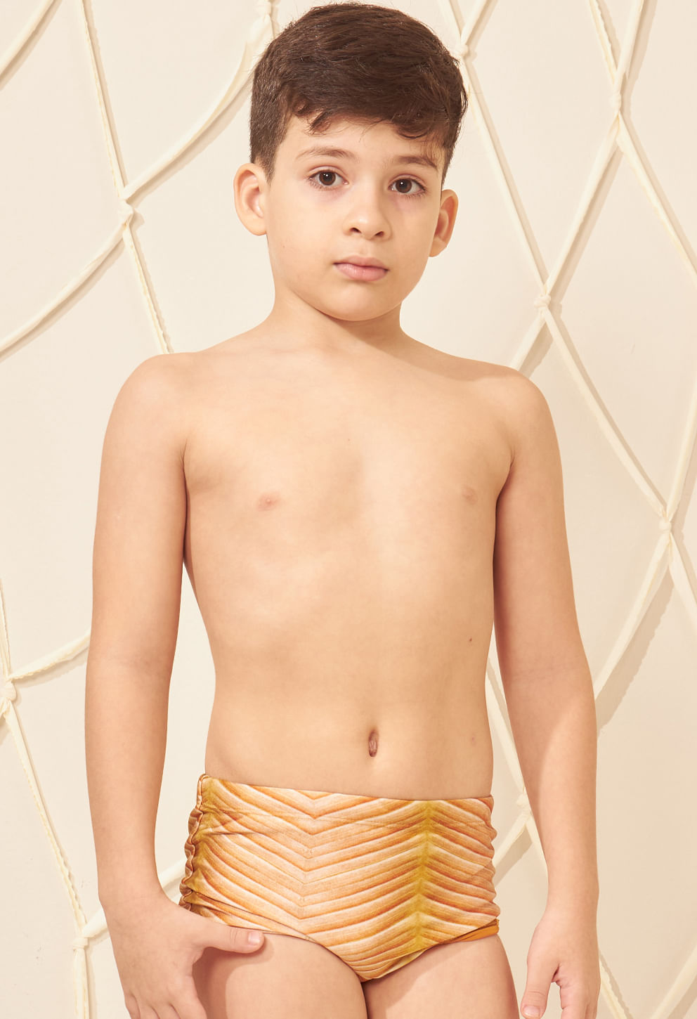 Menino posando com sunga infantil modelo short, padronagem em tons de amarelo, bege e laranja com efeito tridimensional.