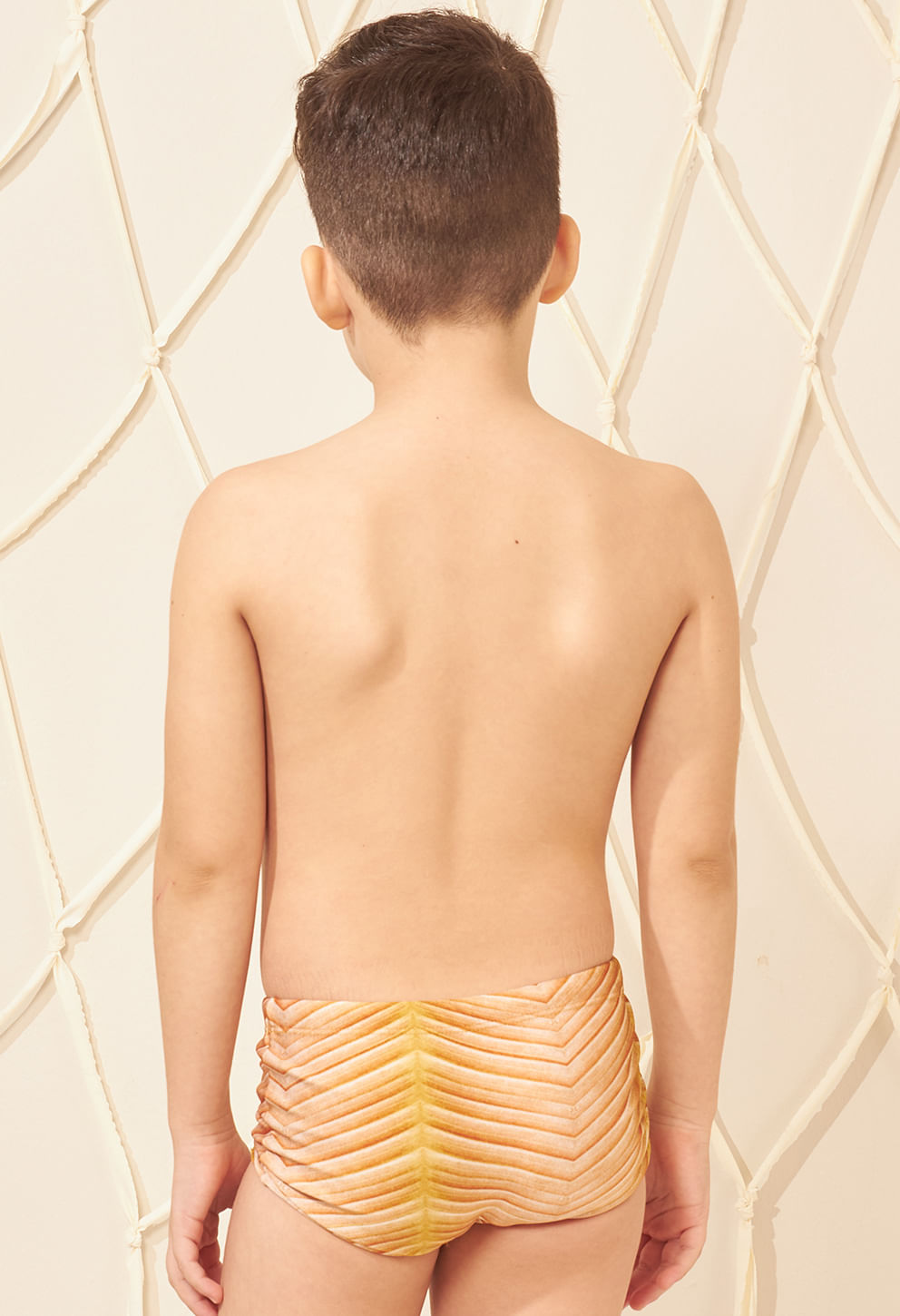 Menino de costas usando sunga infantil short, destacando o ajuste da peça e o padrão em V nas cores amarelo, bege e laranja.