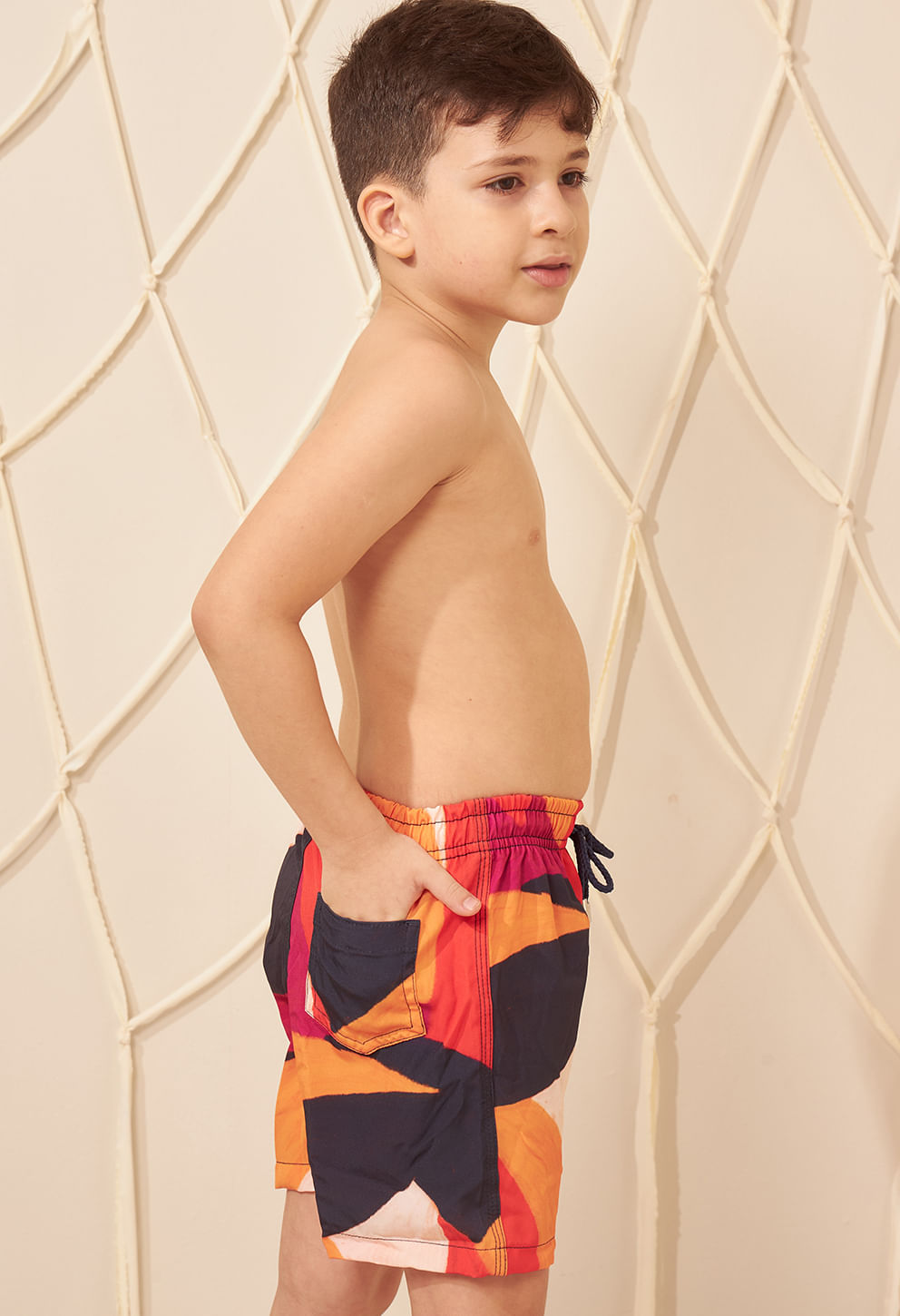 Menino de lado usando bermuda infantil estampada em tons de vermelho, laranja e preto, cós com elástico e cordão azul