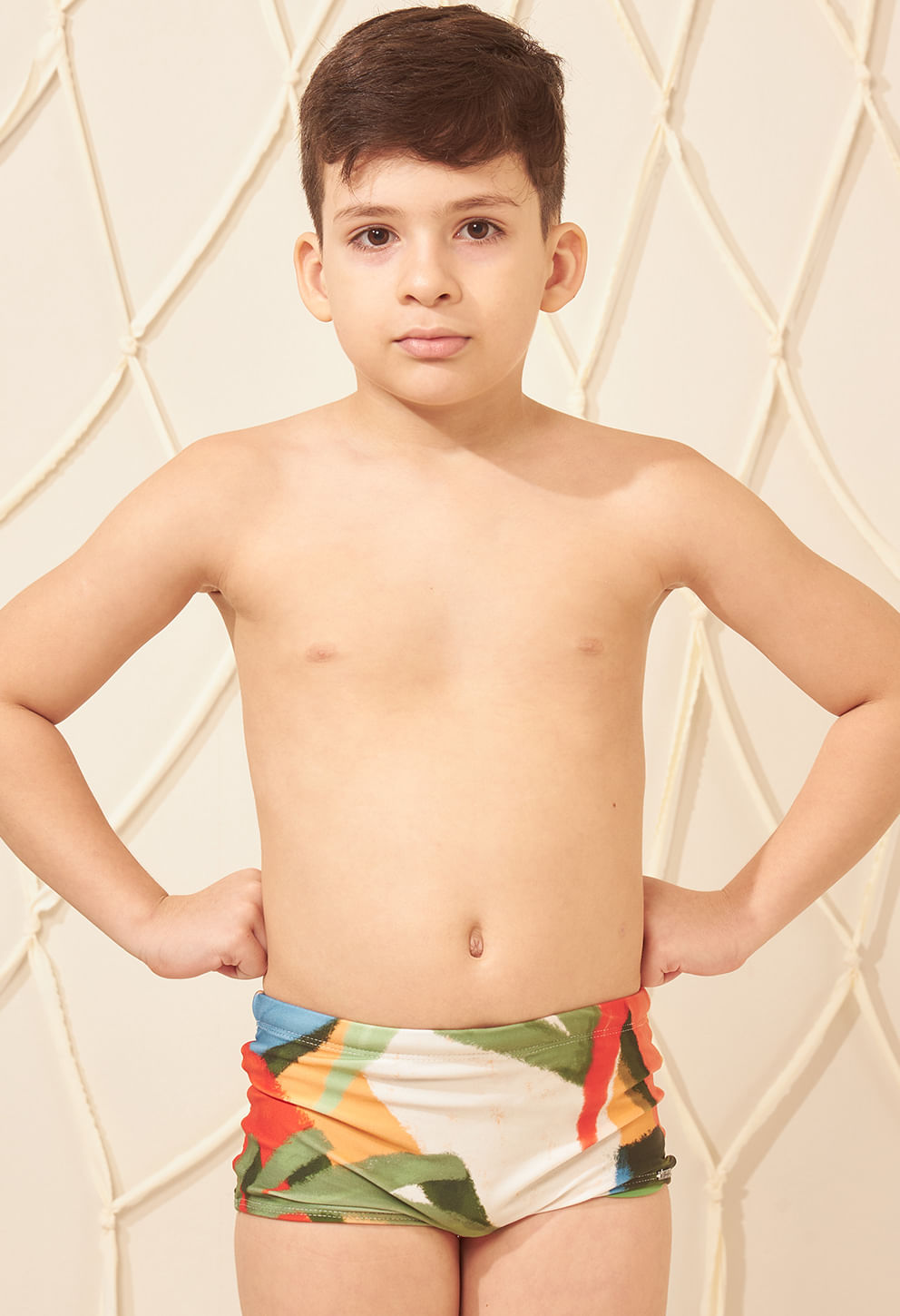 Garoto infantil em pé de frente com as mãos na cintura, vestindo uma sunga short colorida com estampa em tons de verde, laranja, azul e branco.