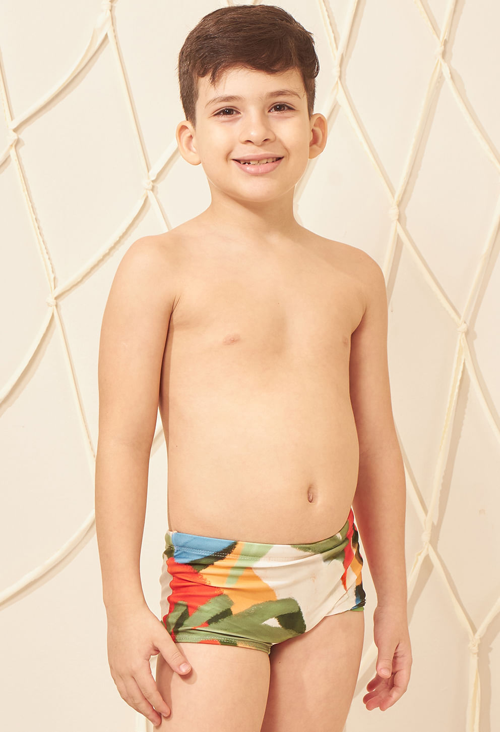 Criança sorridente em pé, mostrando a sunga short infantil estampada com cores vibrantes: verde, laranja, azul e branco.