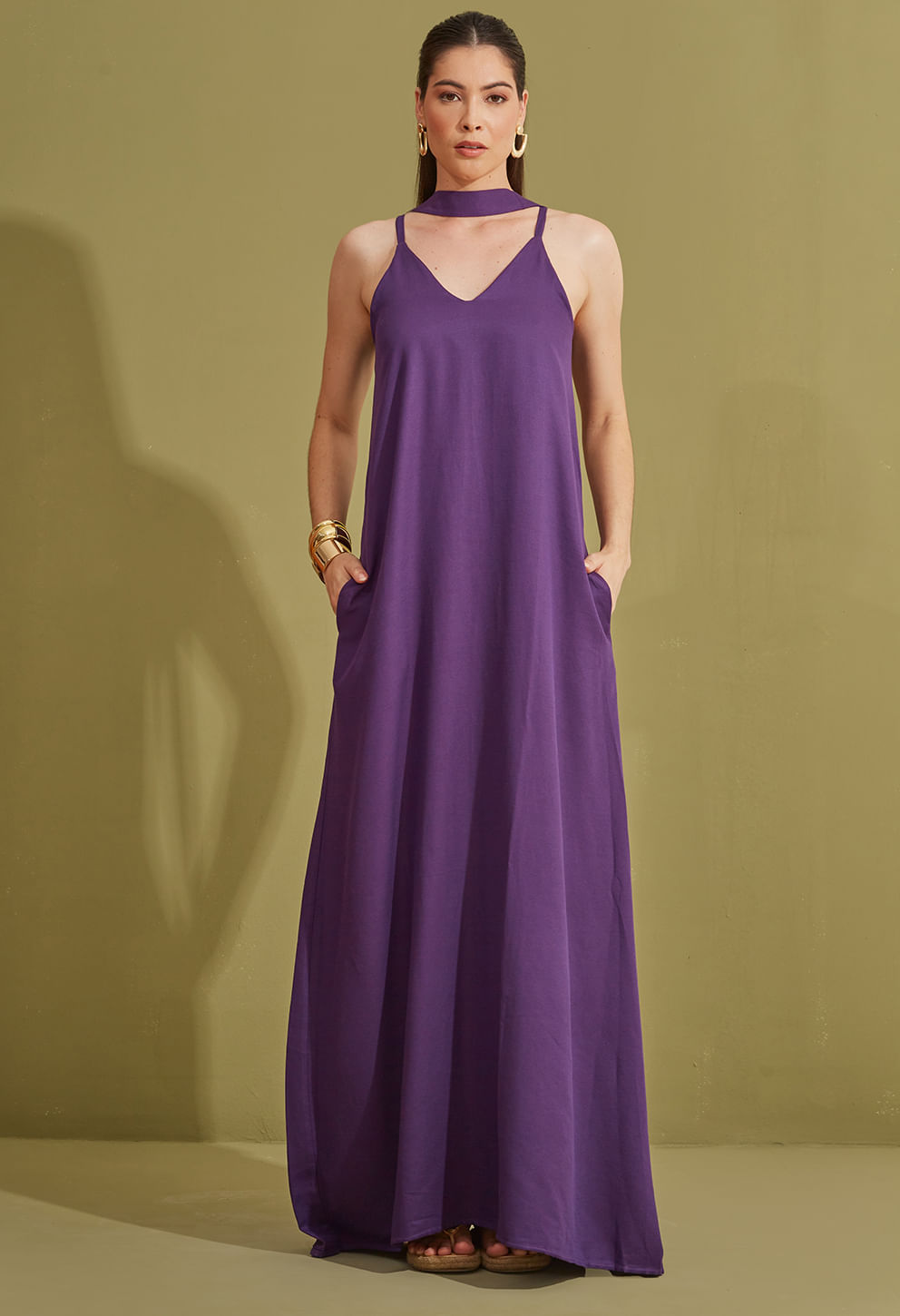 Imagem de vestido longo feminino em tom roxo. Modelo frontal, com vestido de alças finas, decote em V e faixa para amarração na cintura. Tecido fluido e caimento solto. Fundo neutro bege. A modelo usa