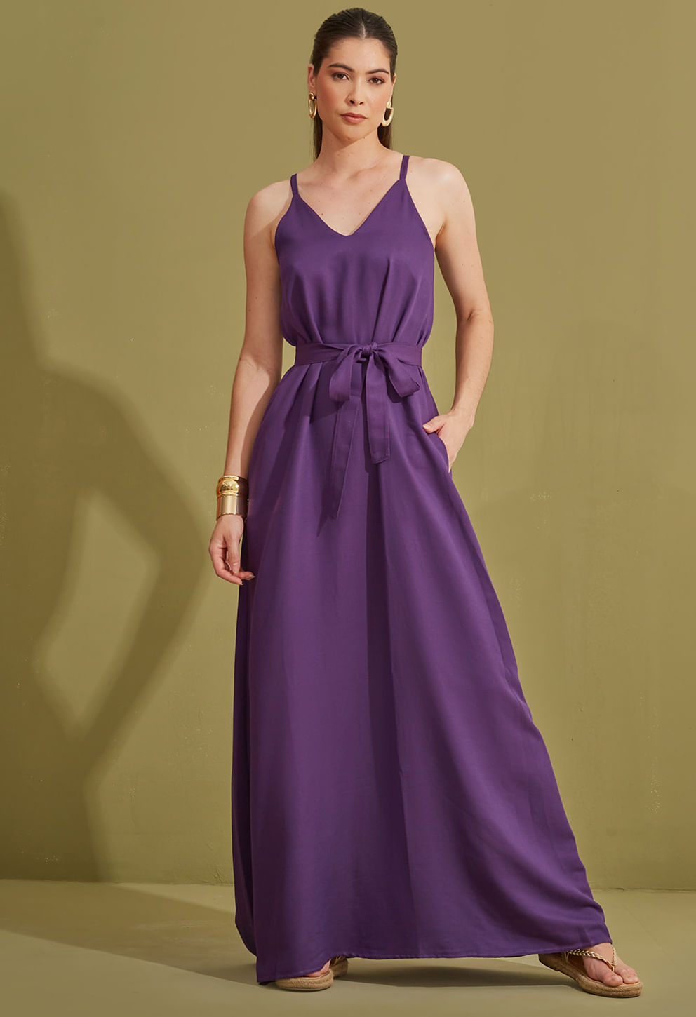 Vestido longo na cor roxa, com alças finas e decote em V, faixa de amarração na cintura destacando a silhueta. Modelo em pose frontal com uma mão no bolso e outra caindo ao lado. Tecido liso