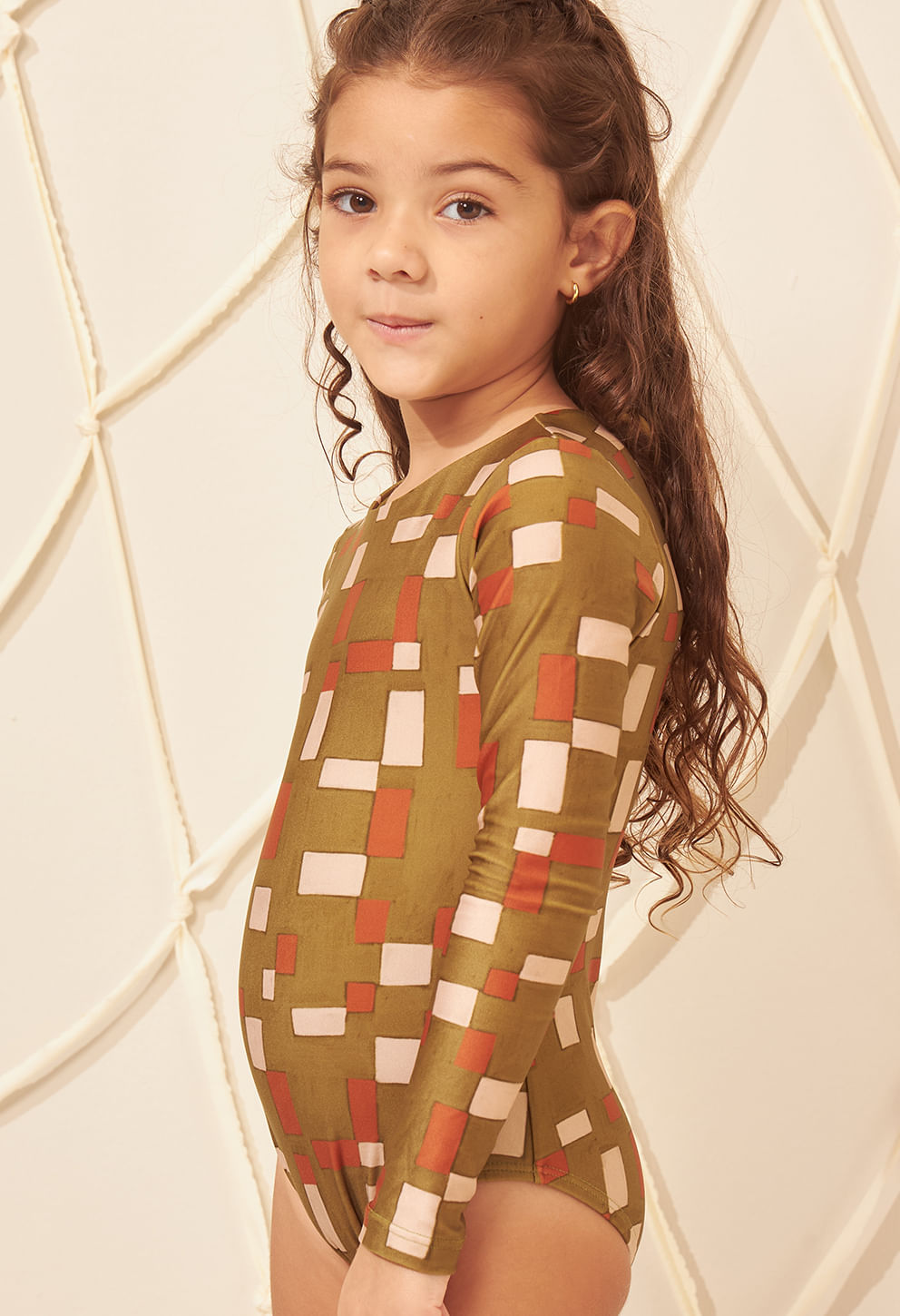 Imagem lateral de menina vestindo maiô infantil manga longa com estampa geométrica verde oliva, laranja e bege