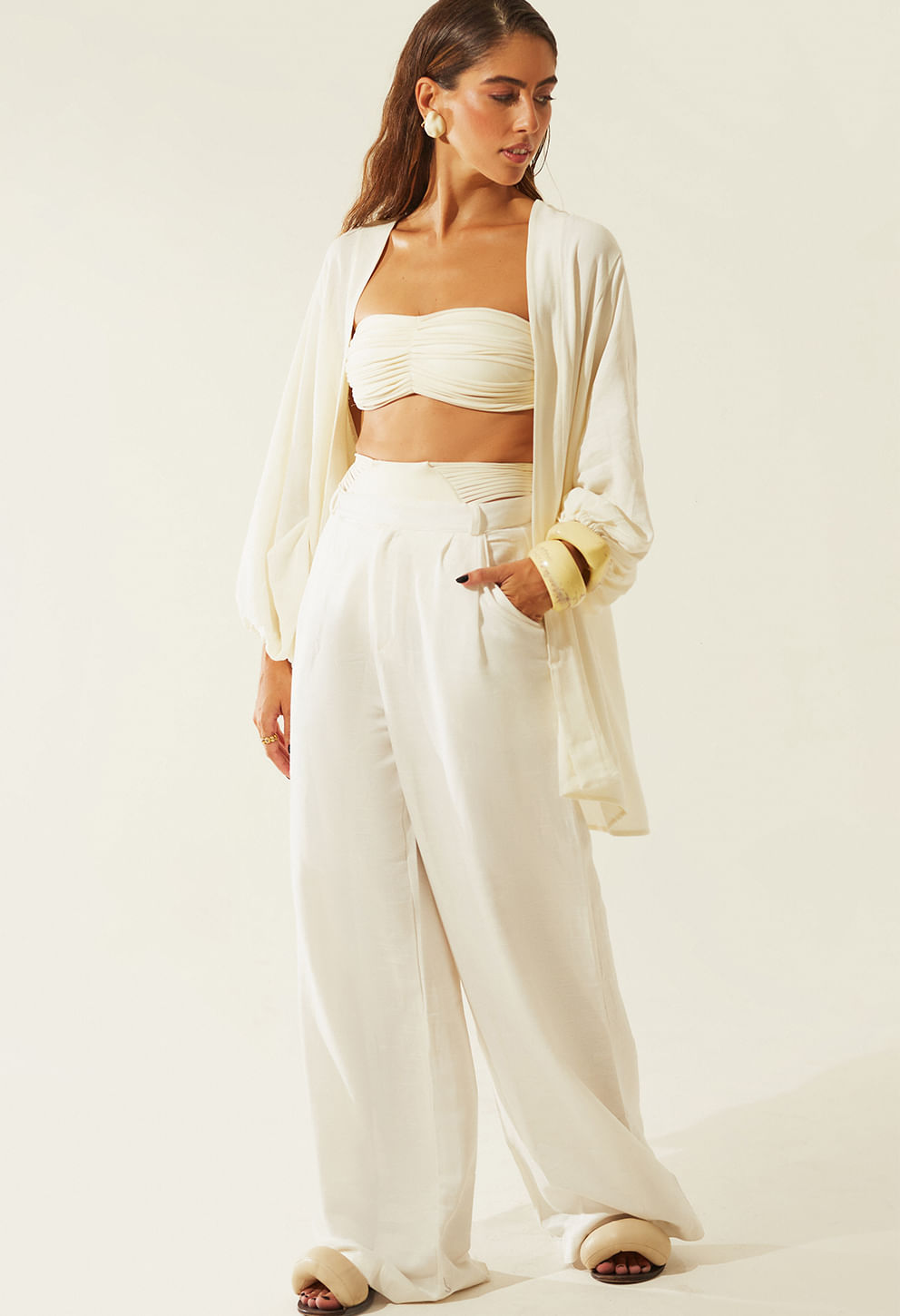 Mulher usando calça pantalona off white de cintura alta com pregas frontais e bolsos laterais, combinada com top faixa franzido