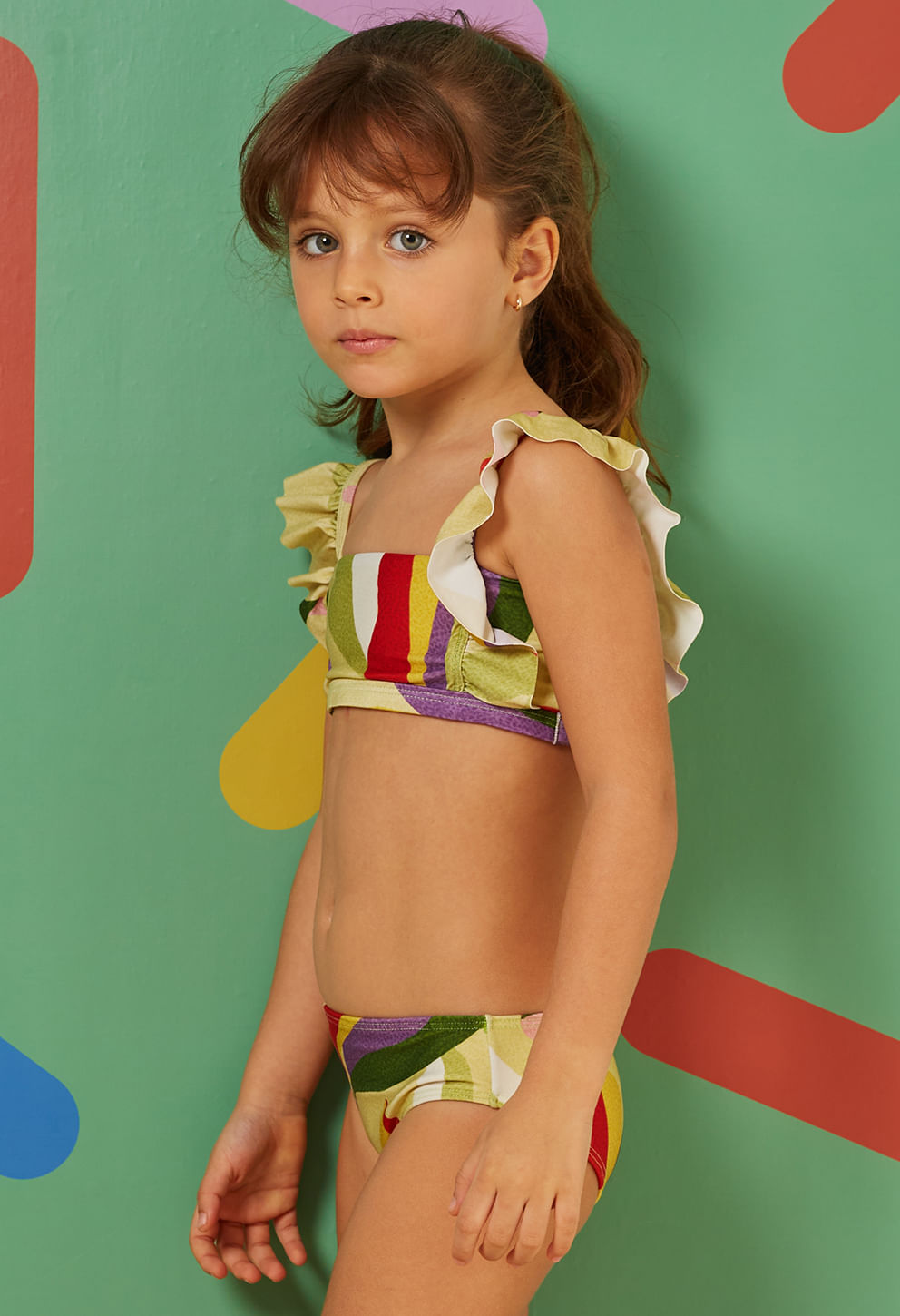 Imagem lateral direita de menina vestindo biquíni infantil com estampa colorida e babados nas alças, fundo verde com detalhes geométricos coloridos.