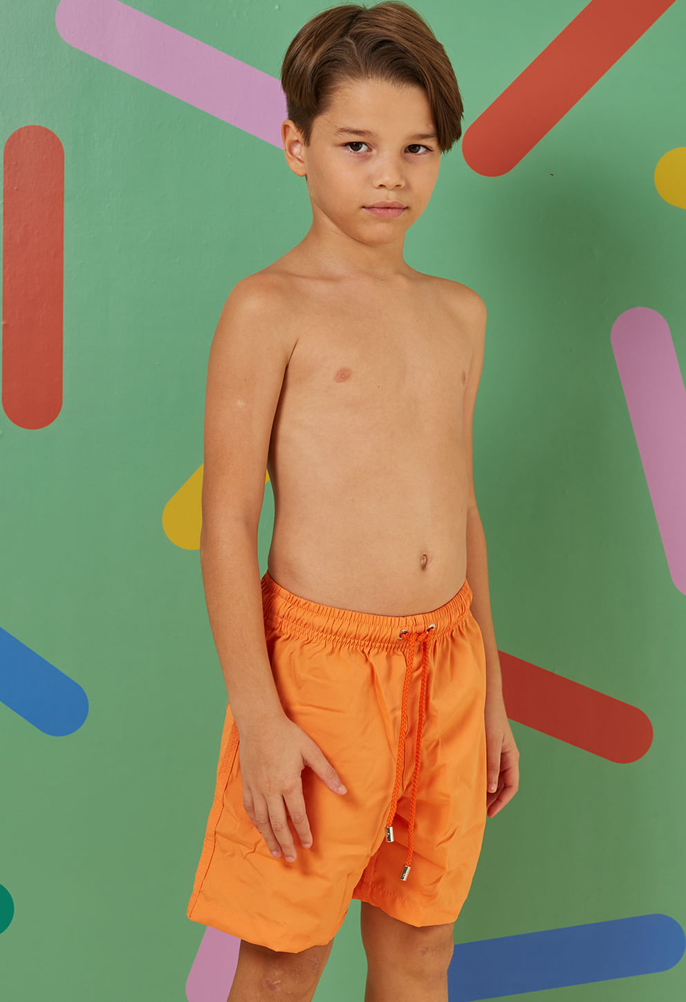 Menino posando com bermuda infantil laranja vibrante, cós com elástico e cordão ajustável, bolsos laterais e traseiro
