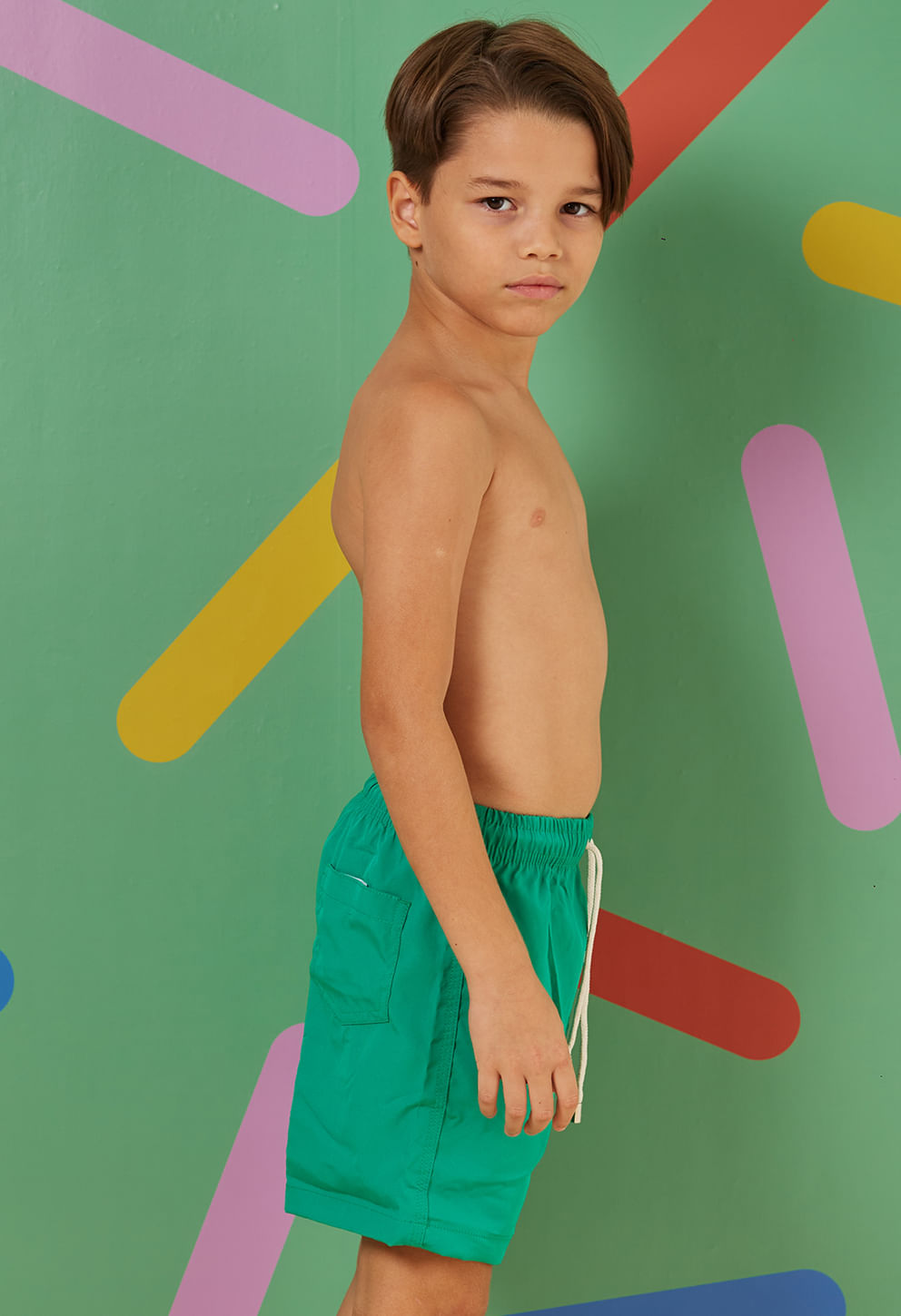 Menino de perfil usando bermuda infantil verde jóia com bolso traseiro e cordão branco