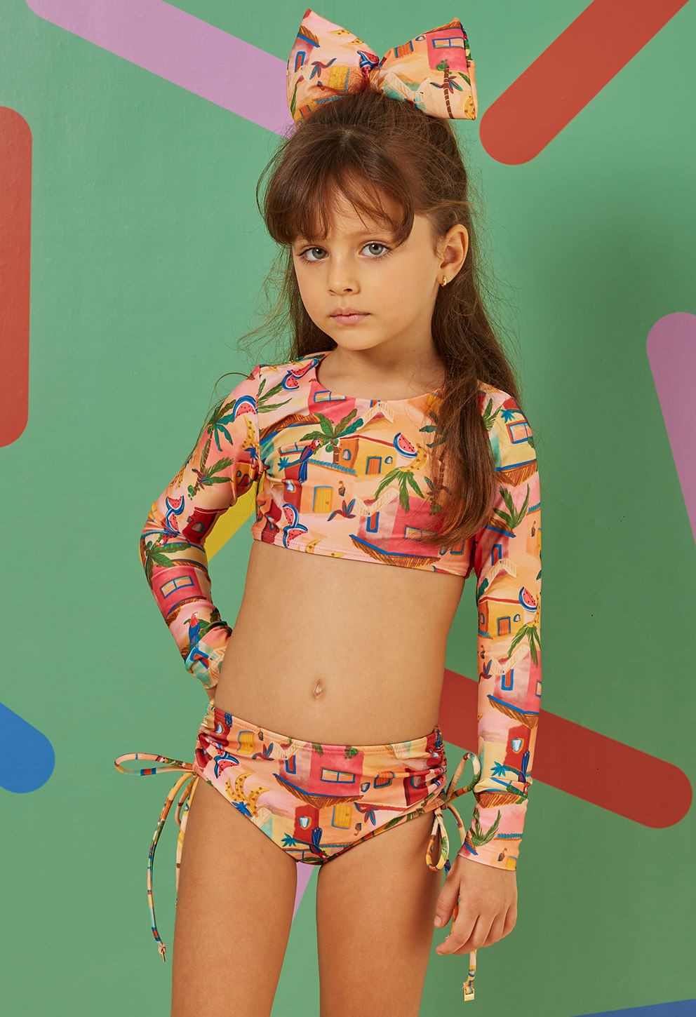 Menina vestindo biquíni infantil Valentina Castelo de Areia com estampa colorida de casas, frutas e coqueiros