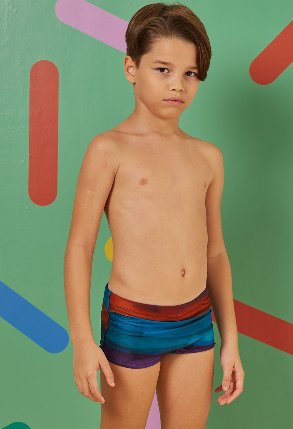 Imagem frontal de um menino usando sunga short infantil listrada em azul, vermelho e roxo