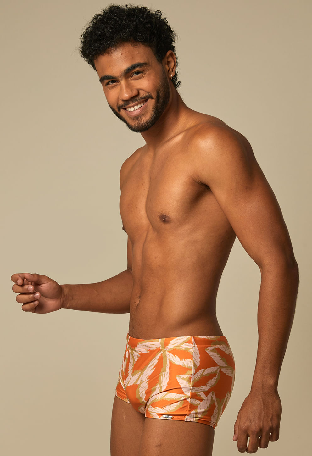 Homem sorridente vestindo sunga short laranja com estampa de folhas, em fundo bege neutro.