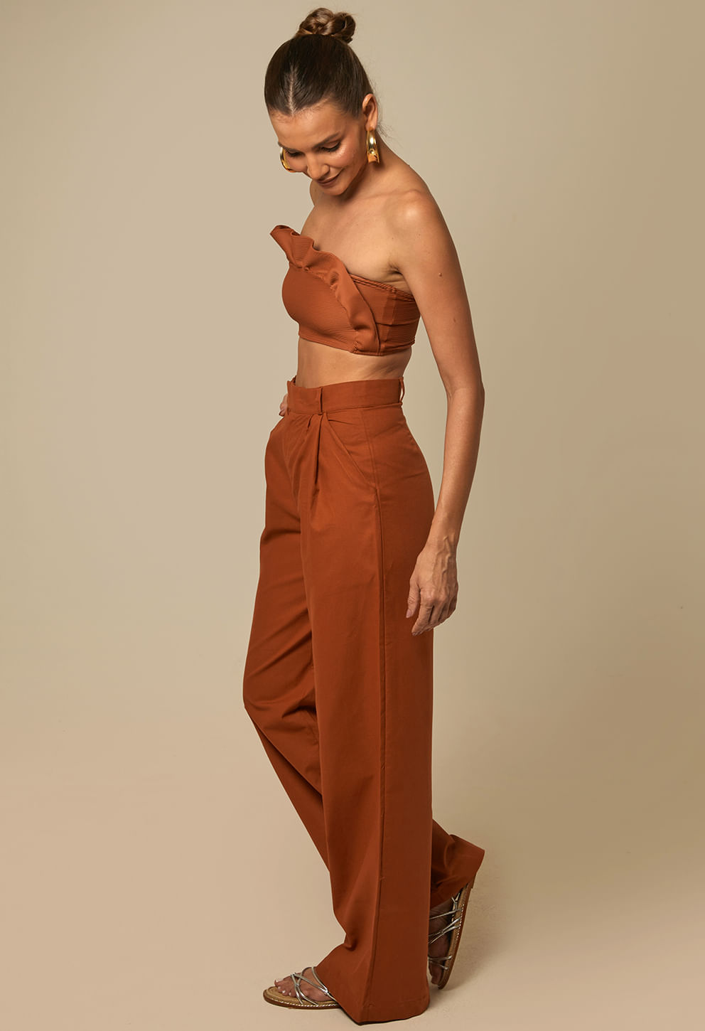 Mulher com calça pantalona terracota de cintura alta, vista de perfil, posando com a mão na cintura.