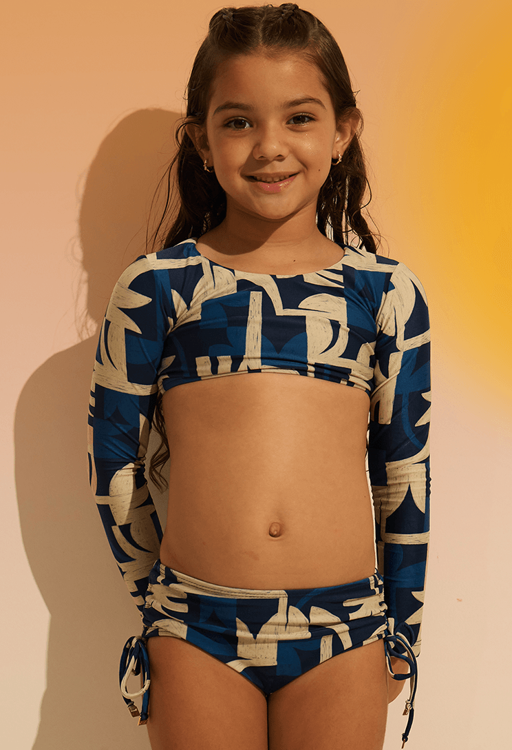Menina sorridente vestindo biquíni Valentina Costeira azul e bege, top cropped de manga longa e calcinha com amarração lateral