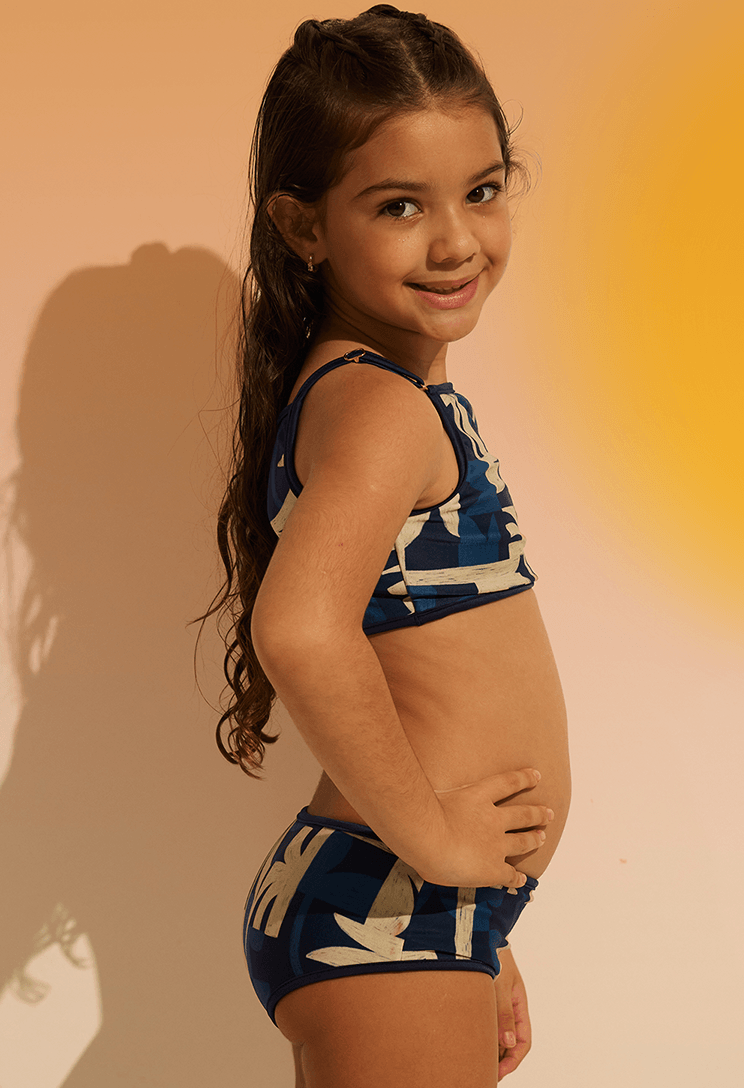 Imagem lateral de menina sorridente usando biquíni Bebela Costeira com estampa abstrata azul e bege