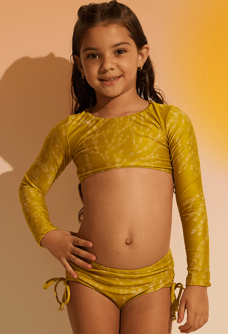 Menina vestindo biquíni Valentina Corais amarelo mostarda com estampa branca, blusa de manga longa e calcinha com amarrações